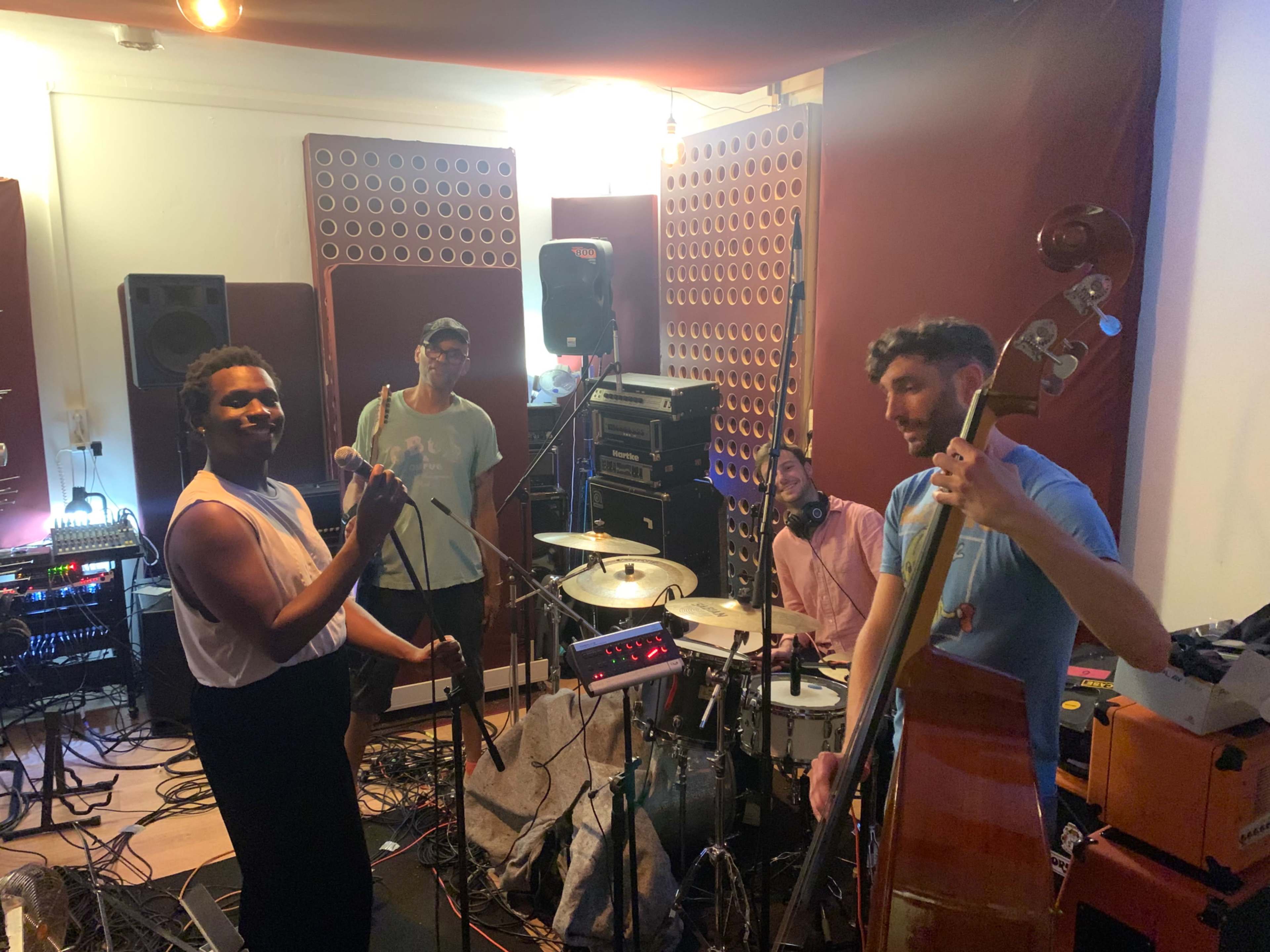 A group of four musicians is playing instruments in a recording studio, surrounded by sound equipment and acoustic panels.