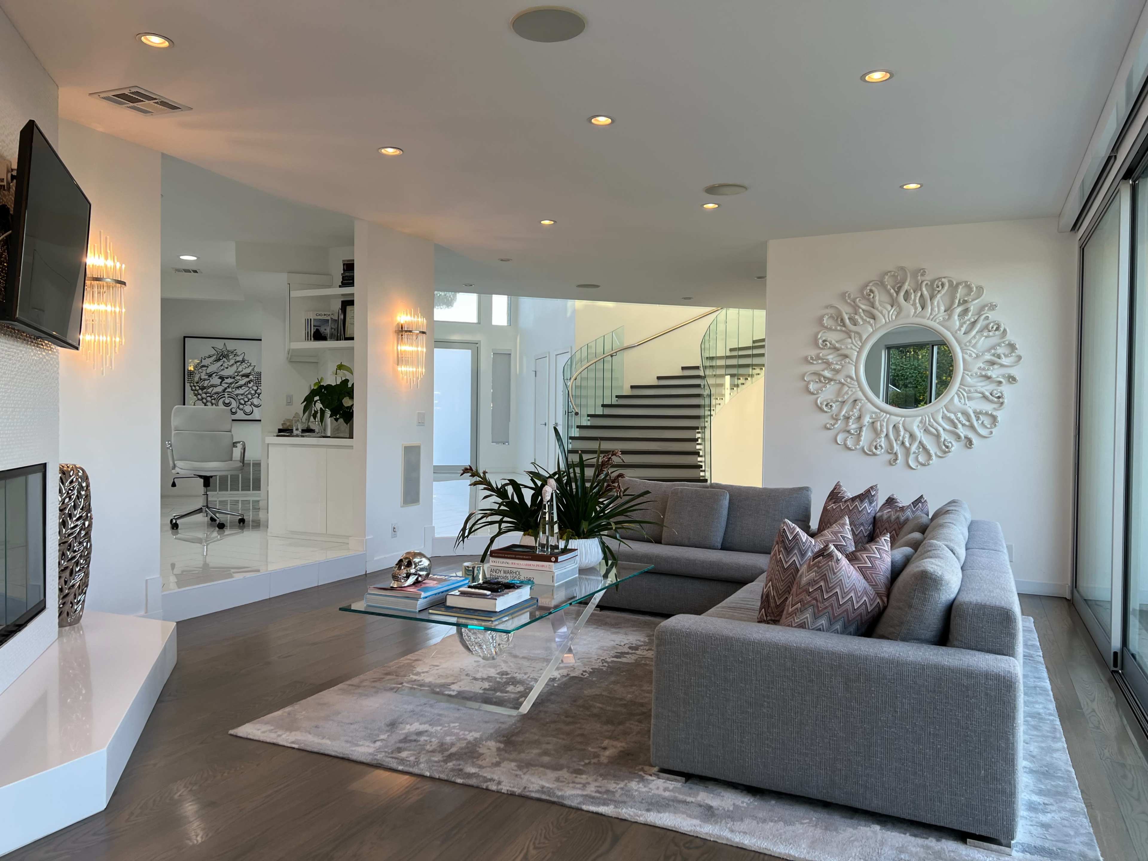 A modern living room featuring a gray sectional sofa, a glass coffee table on a light-colored rug, and large windows with a view of a staircase.