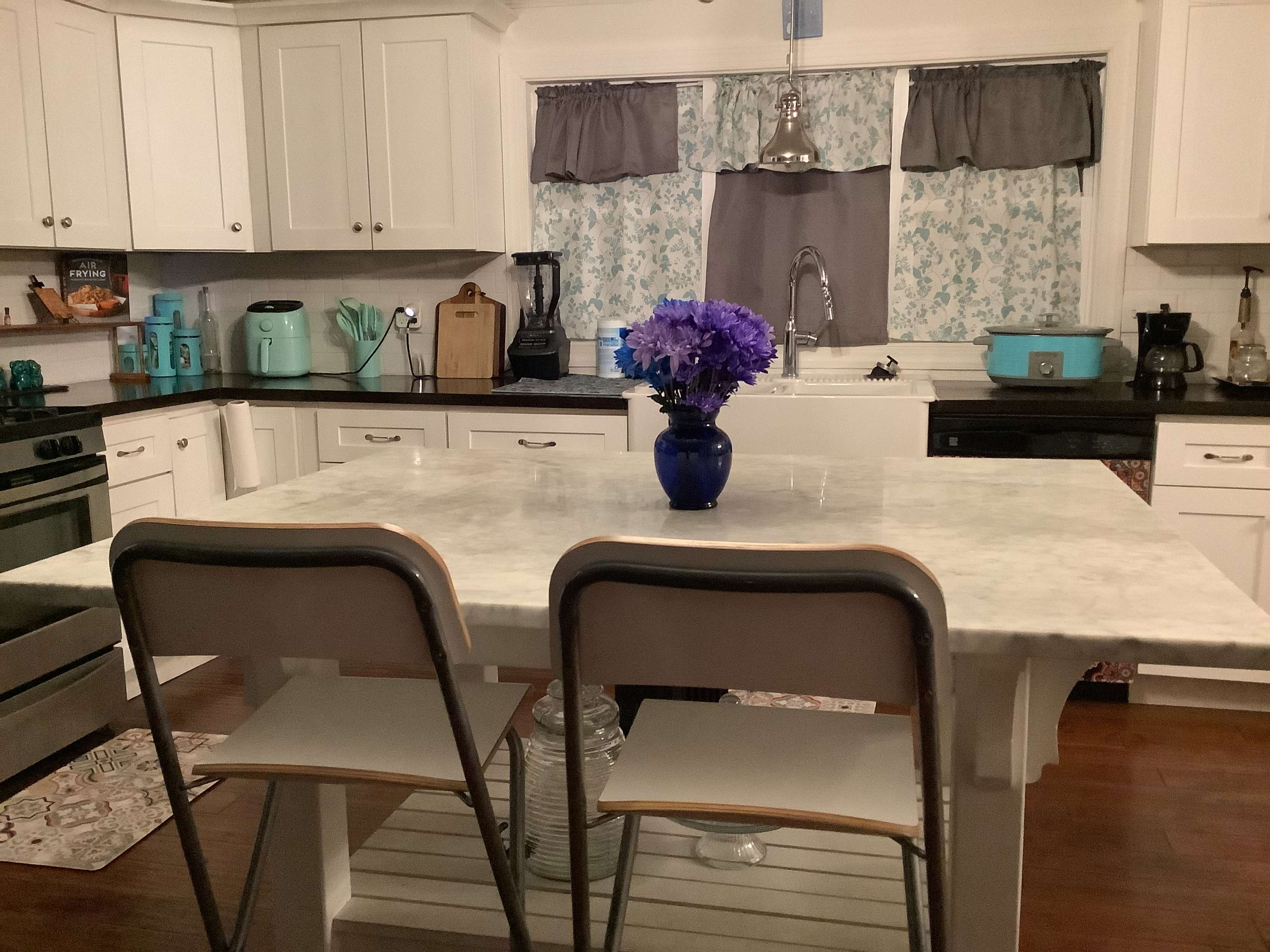 A kitchen featuring a marble-topped island with two chairs and a vase of purple flowers, surrounded by white cabinets and appliances.