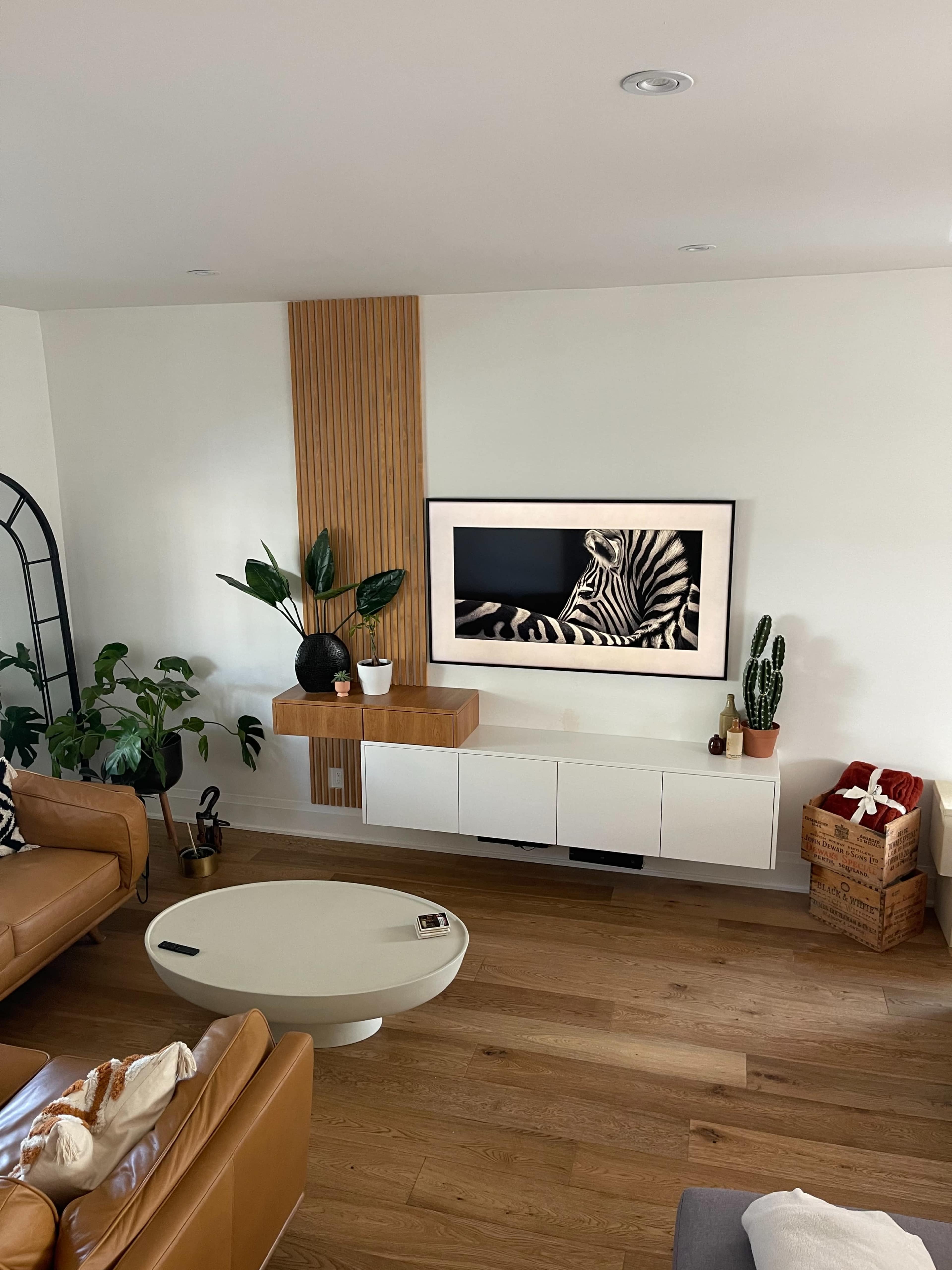 A modern living room featuring a wall-mounted television above a white cabinet, surrounded by indoor plants and a round coffee table on a wooden floor.