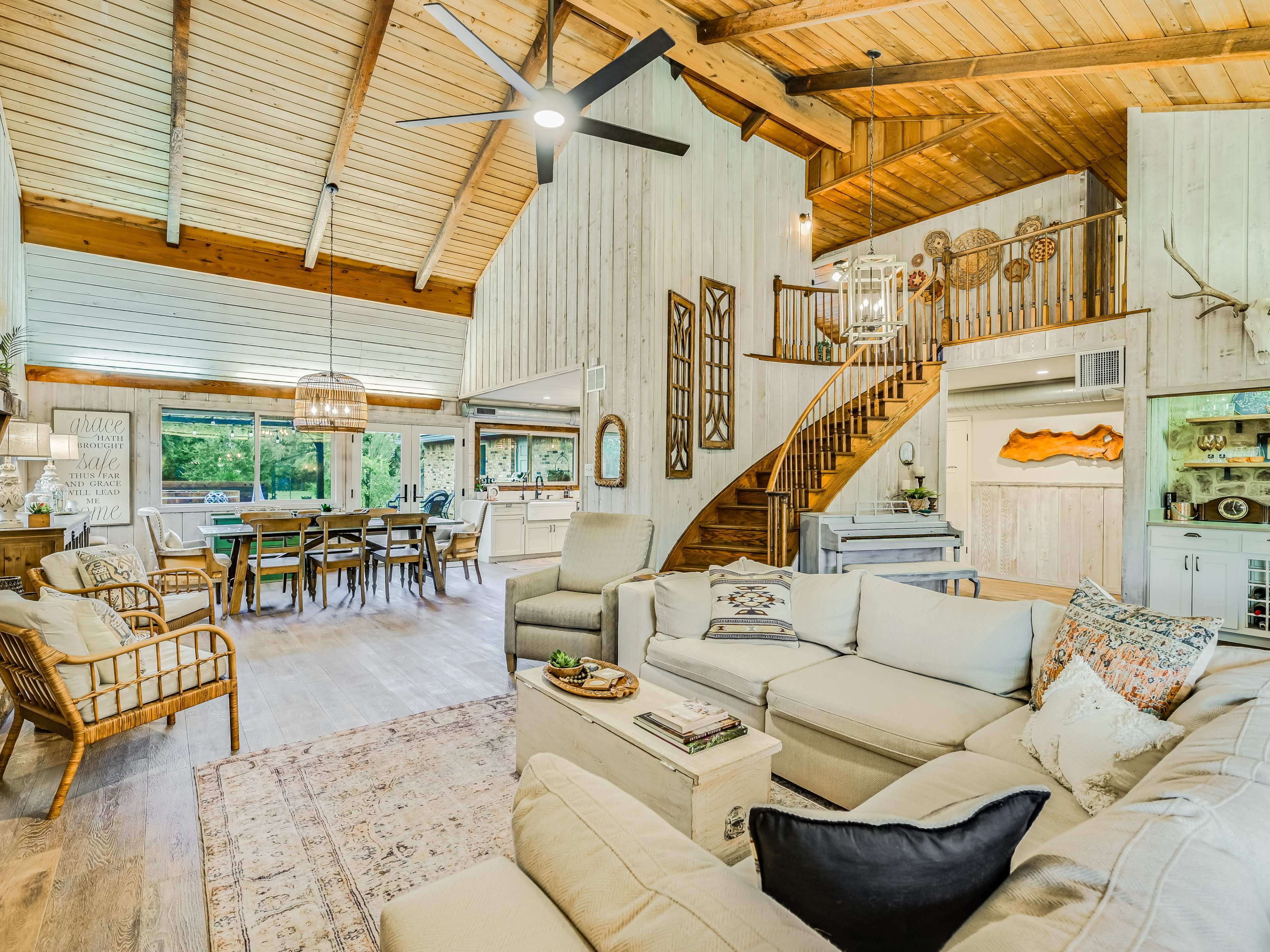 A spacious, open-concept living area features a white sectional sofa, large windows, wooden beams, and a staircase leading to an upper level.