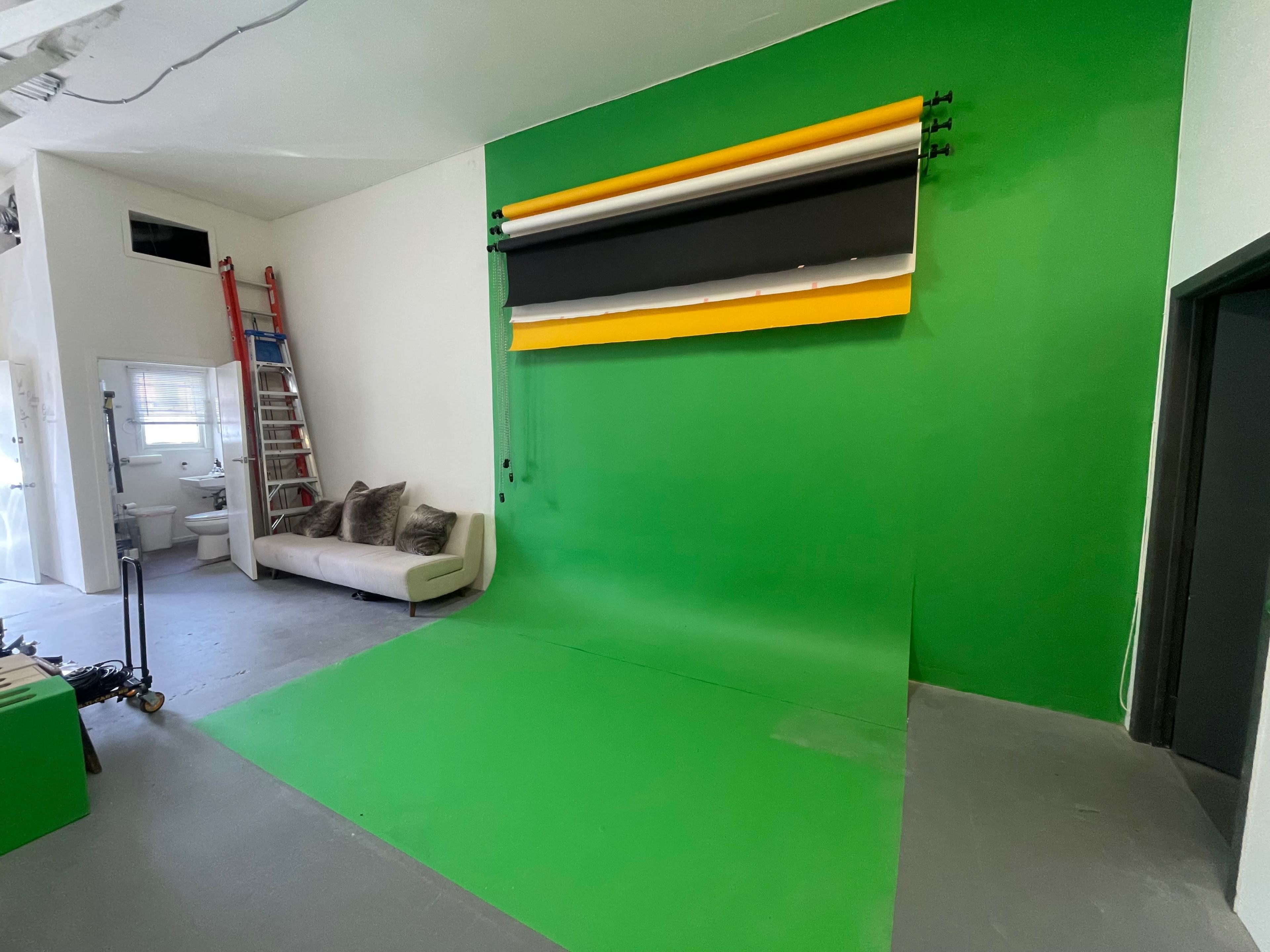 A studio space with a green backdrop, a white wall, a sofa, and a ladder leaning against the wall.