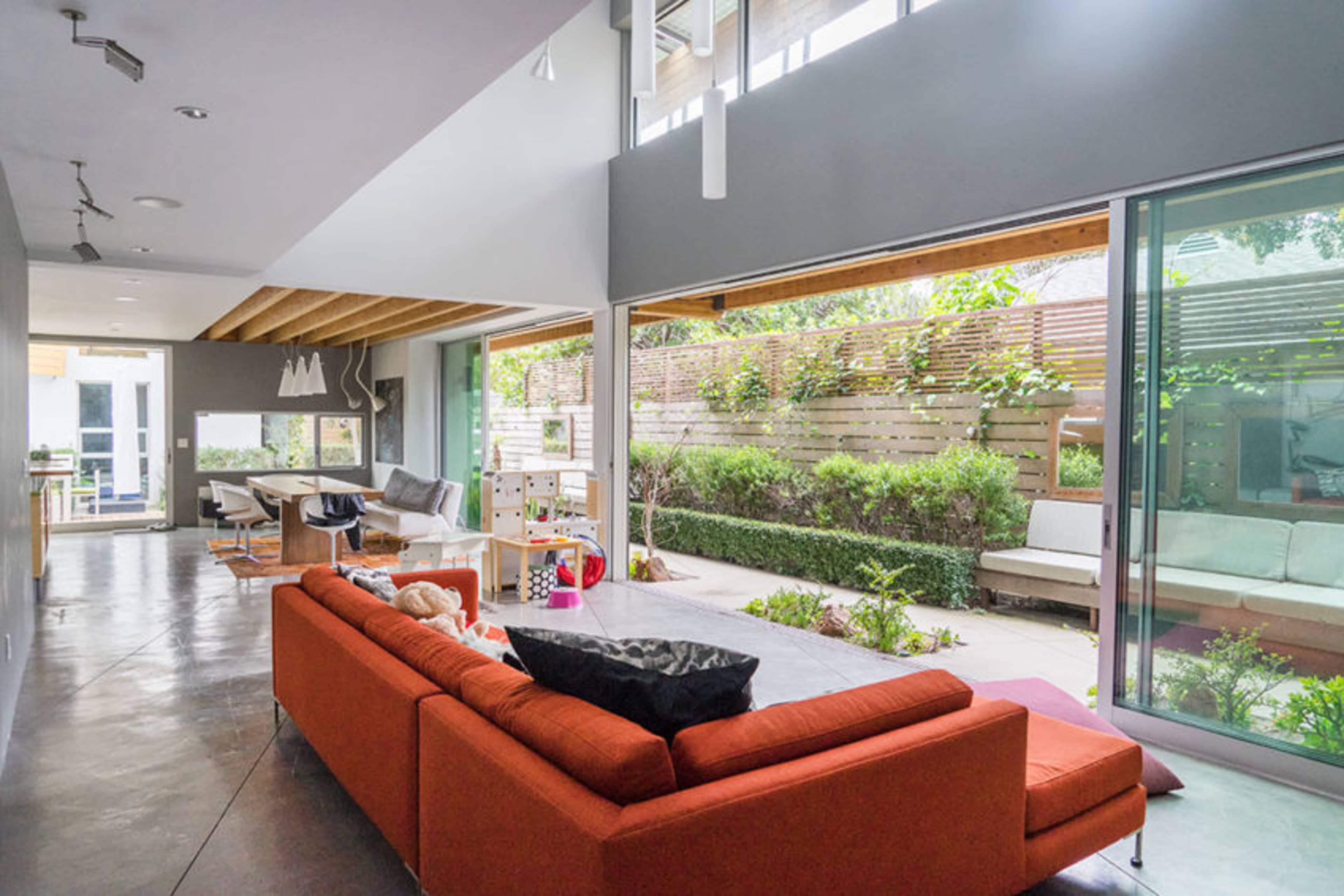 The image shows a modern living space with an open layout, featuring a large orange sofa, a dining area, and large sliding glass doors that open to a green garden.