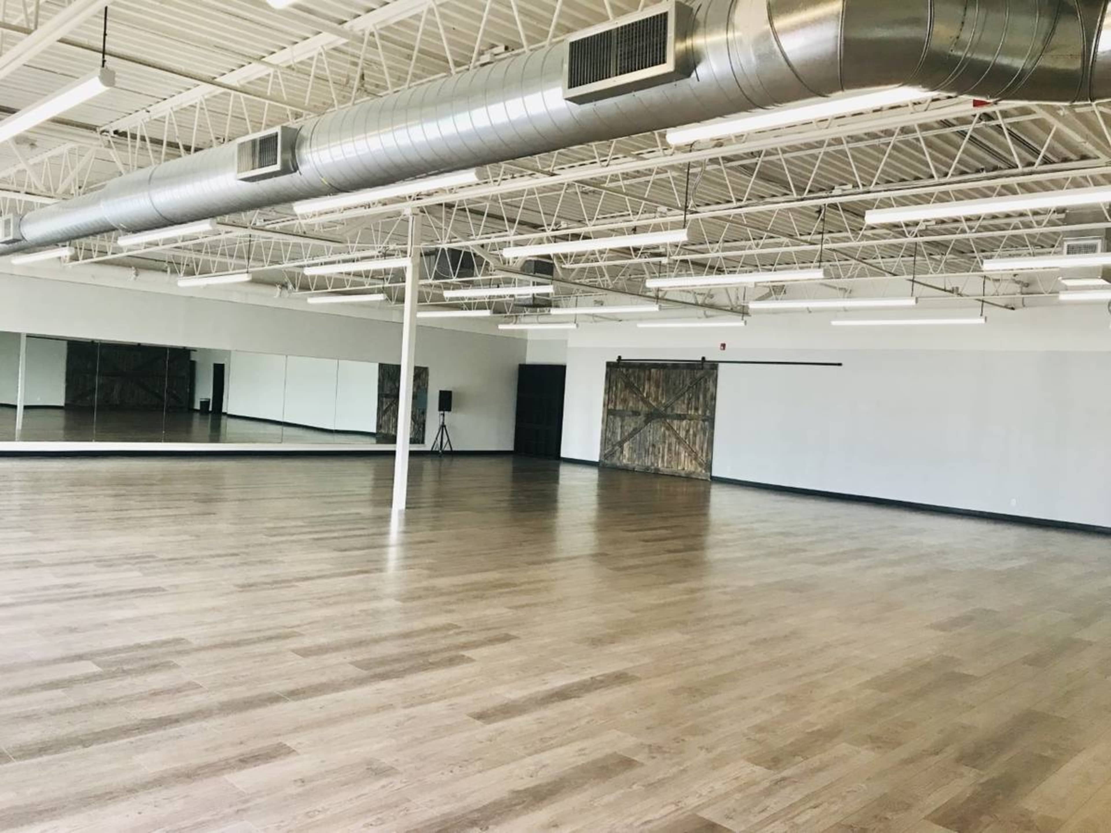 An empty dance studio with wooden flooring, large mirrors on one wall, and industrial-style metal ductwork overhead.