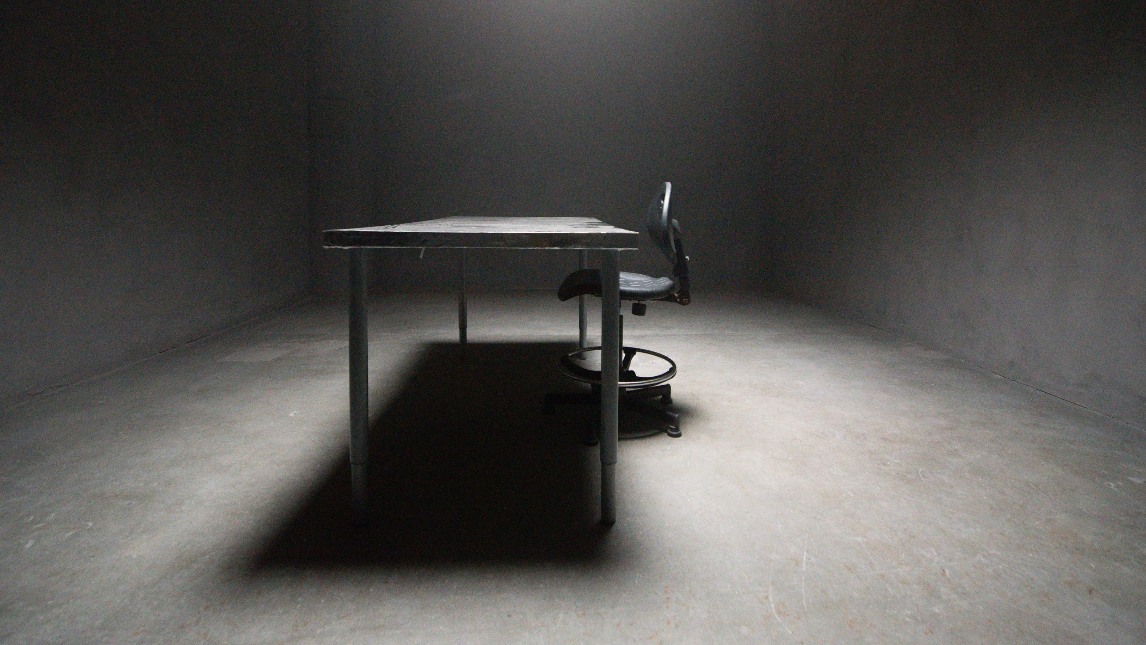 A solitary table and a chair are positioned in a dimly lit, empty room with gray walls.
