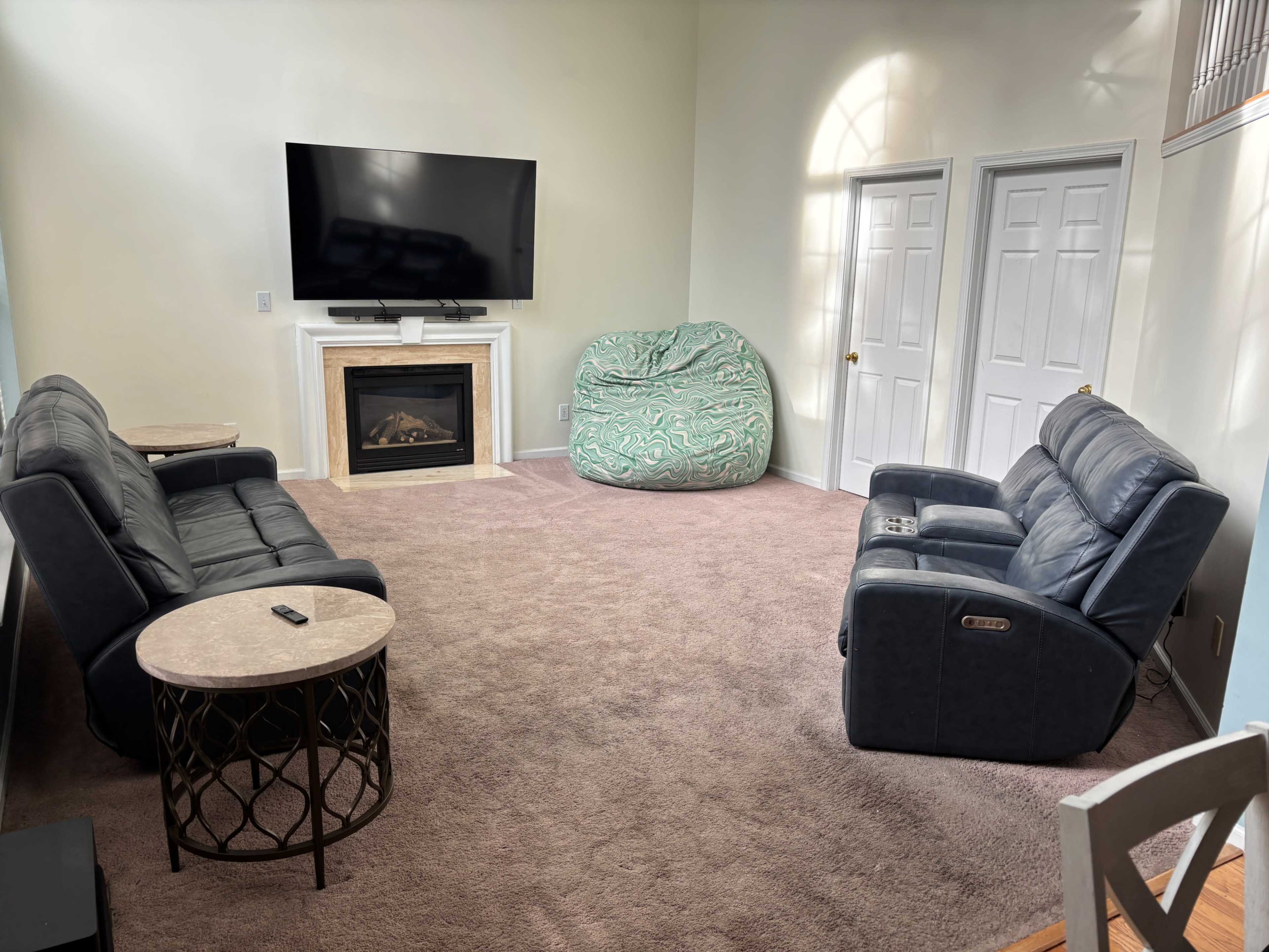 The image shows a spacious living room with two leather sofas facing a large television, a round table, a bean bag chair, and a fireplace, all on plush carpeting.