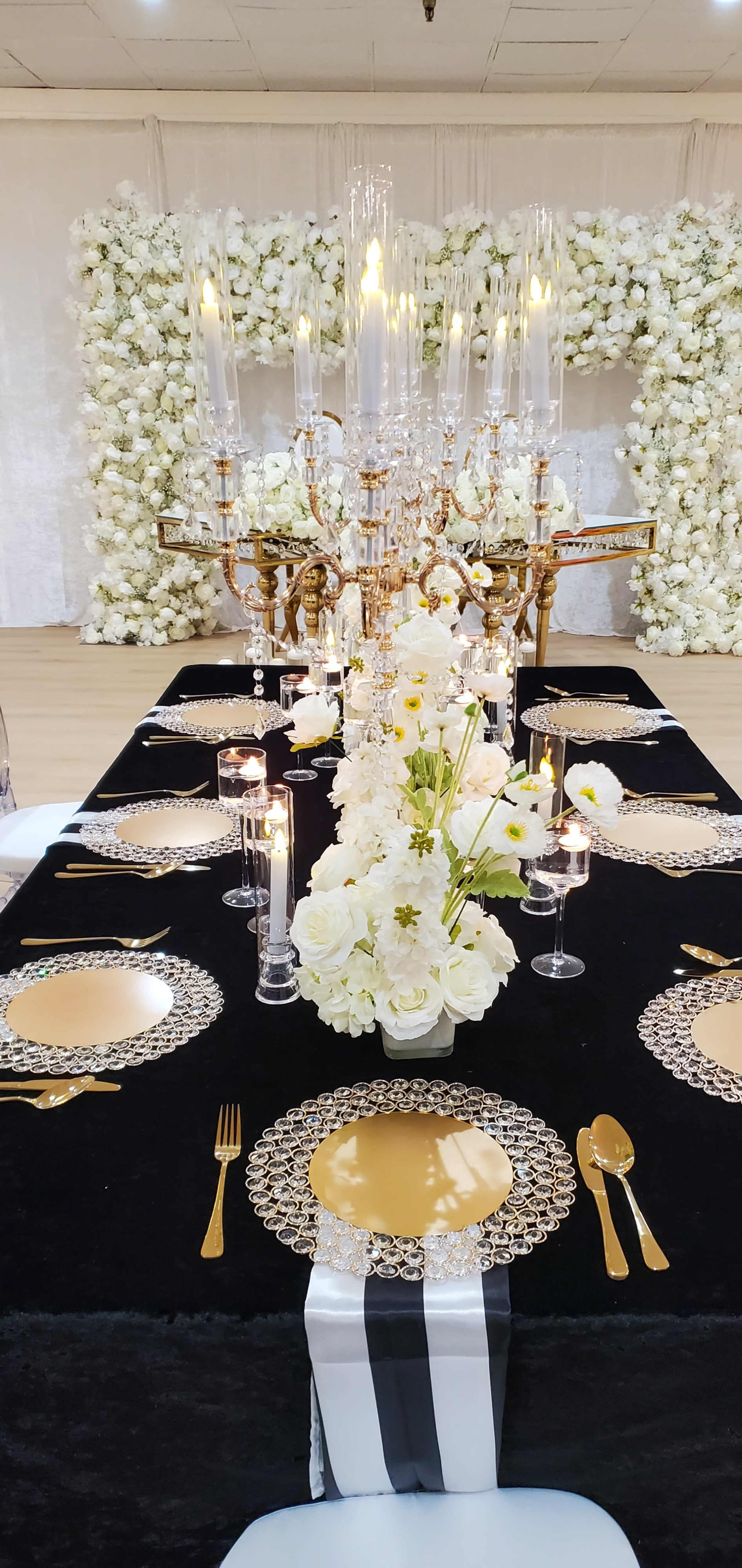 The image shows a long banquet table adorned with elegant place settings, a floral centerpiece, and a decorative candelabrum amidst a lavish backdrop of white flowers.