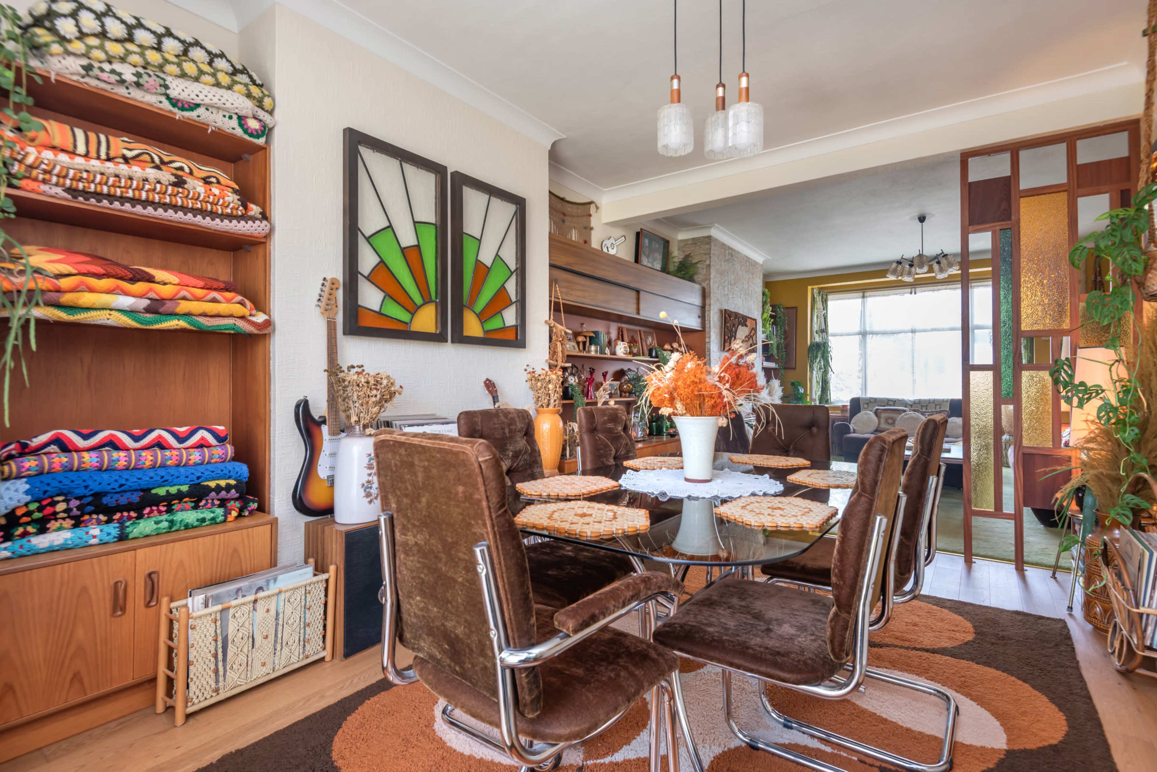 A dining area features a round glass table surrounded by brown upholstered chairs, with colorful blankets displayed on shelves and decorative items accentuating the space.