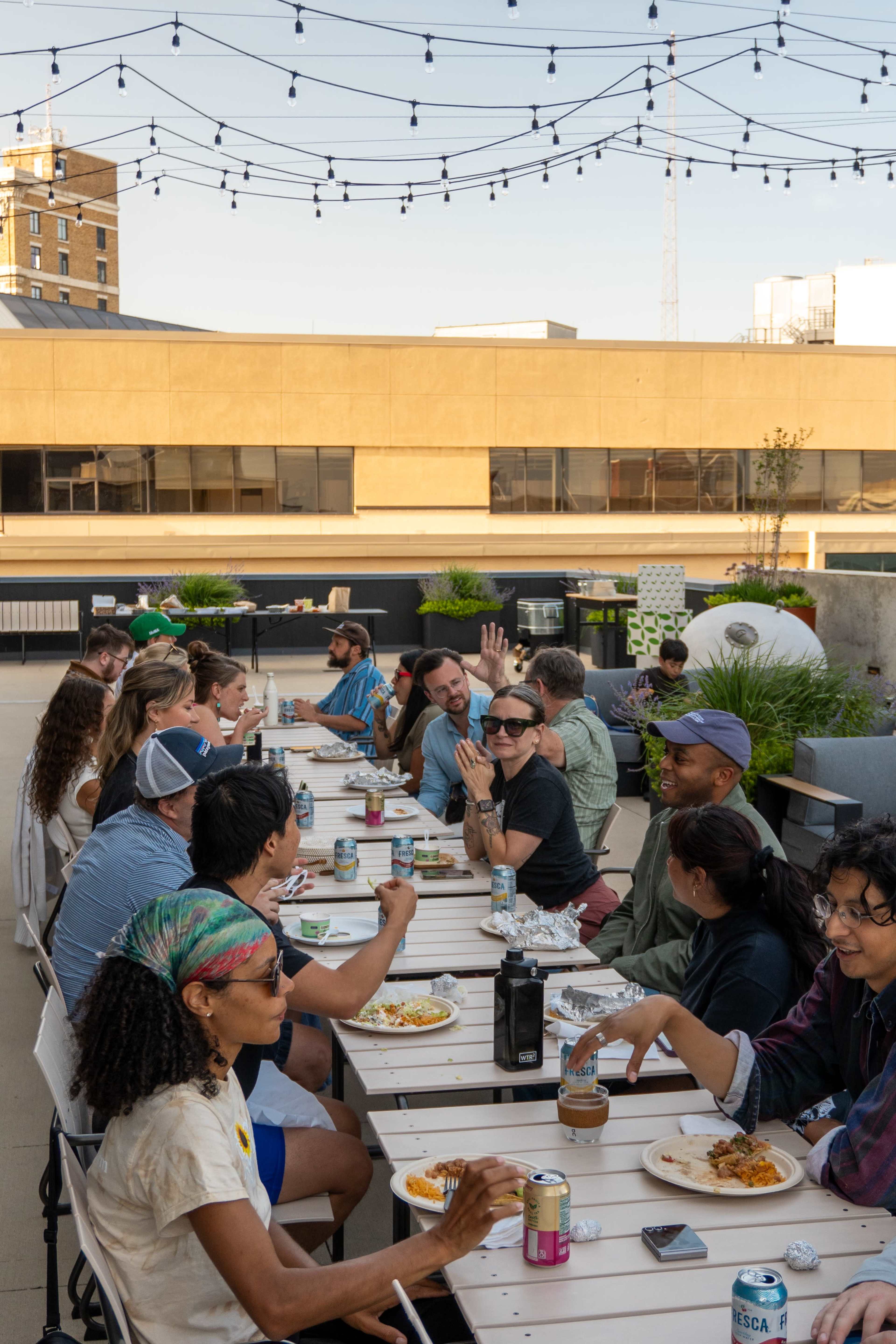 A group of people sits at a long table outdoors, sharing food and drinks under string lights in an urban setting.
