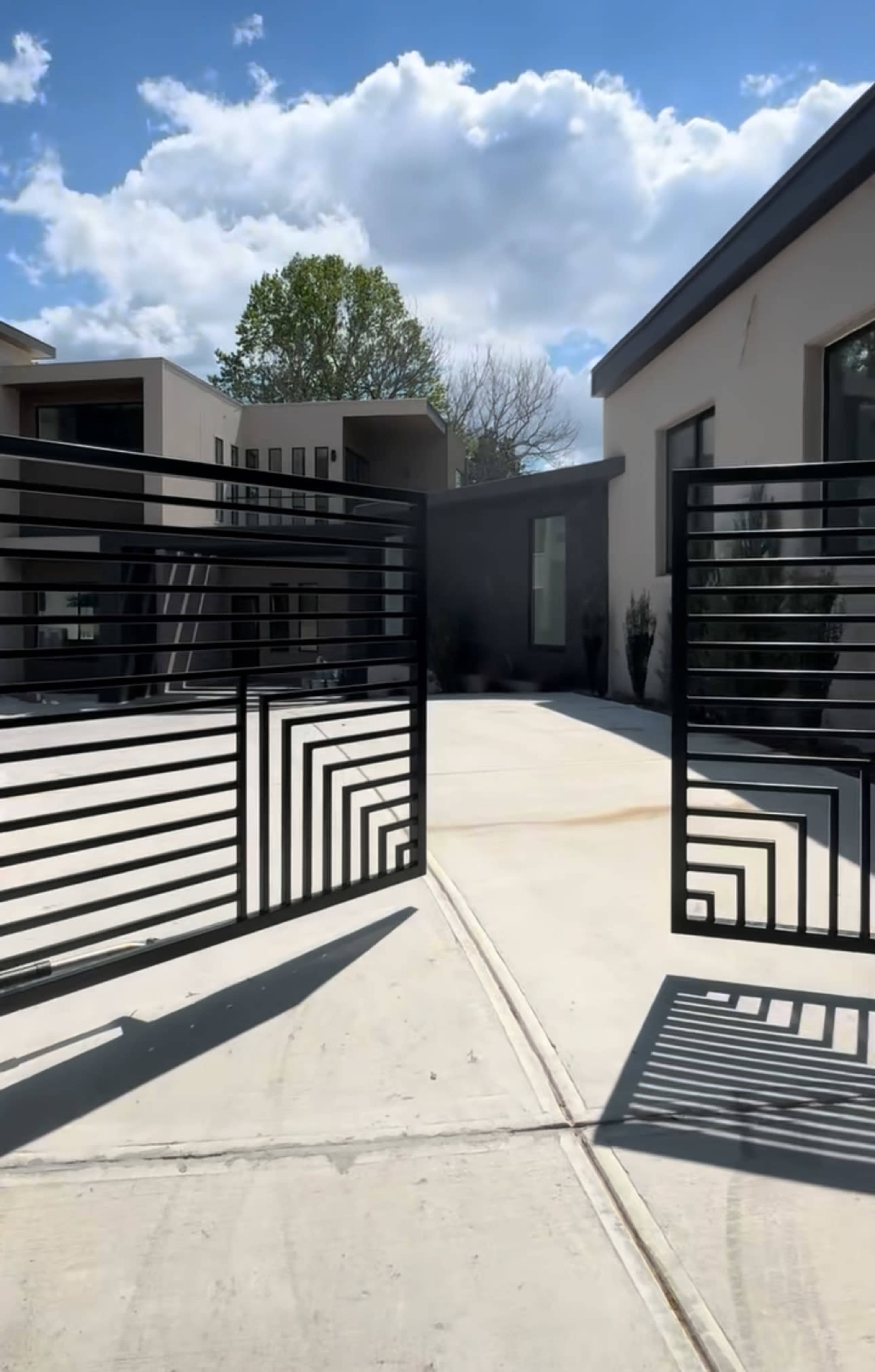Two black metal gates stand open, leading to a modern courtyard with landscaped greenery and a light-colored concrete pathway.