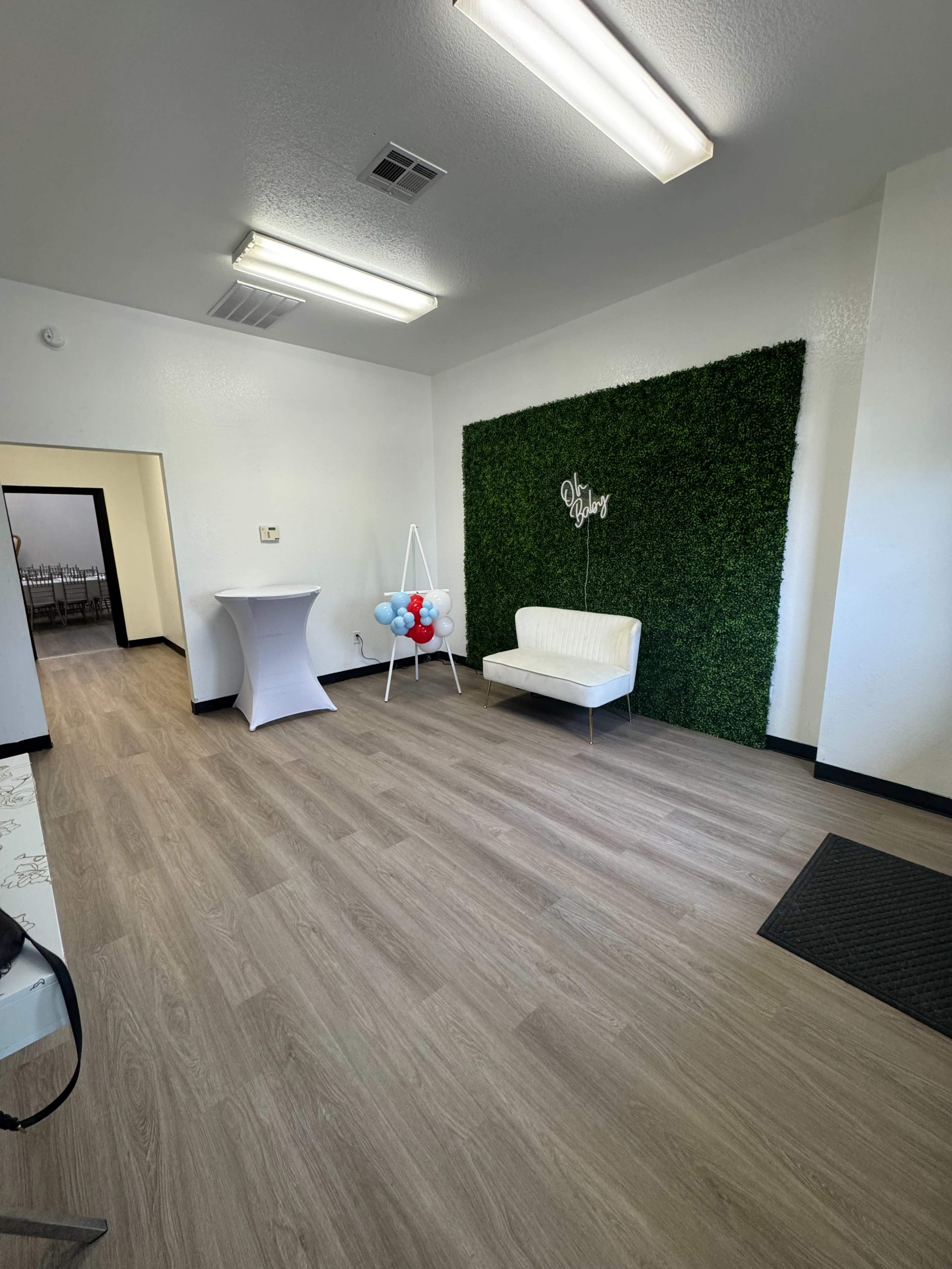 The image shows a spacious, empty room with a green plant wall, a white couch, a tall table, and a sign on the wall.