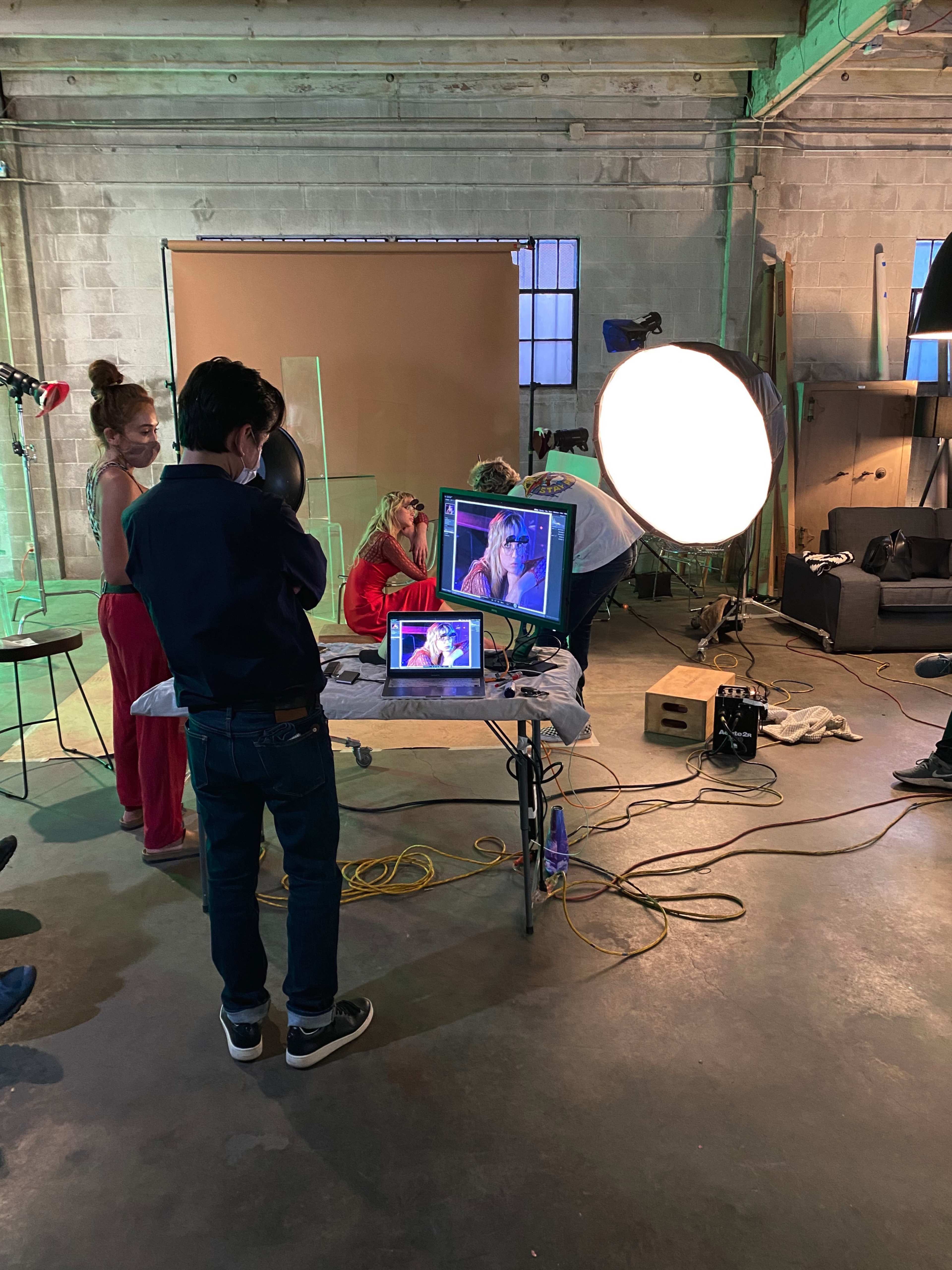 A group of individuals is working in a studio, with one person monitoring a video feed on a laptop while others are positioned in front of a large softbox light and a backdrop.