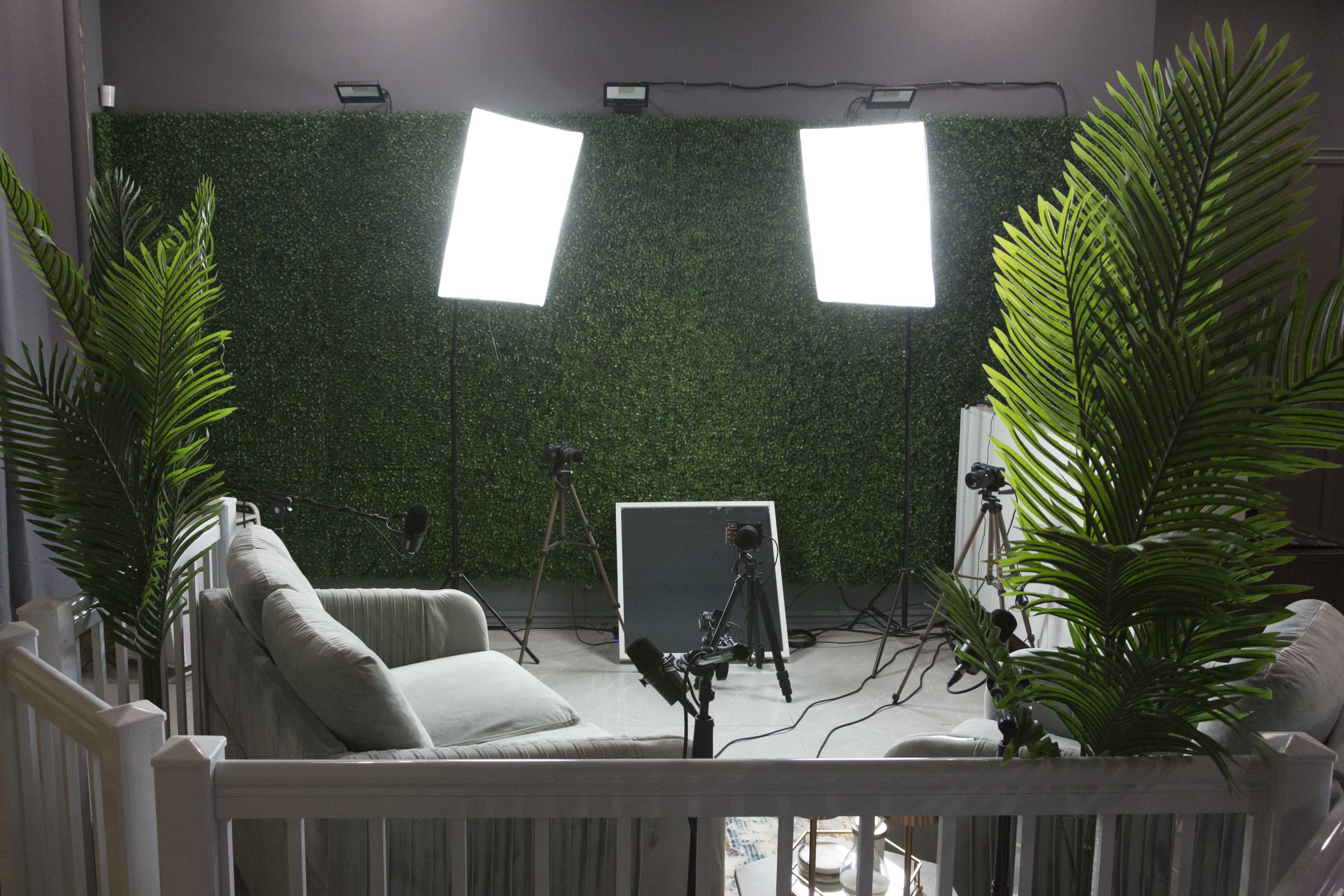 A well-lit studio setup features a green foliage backdrop, a sofa, and cameras positioned for recording.