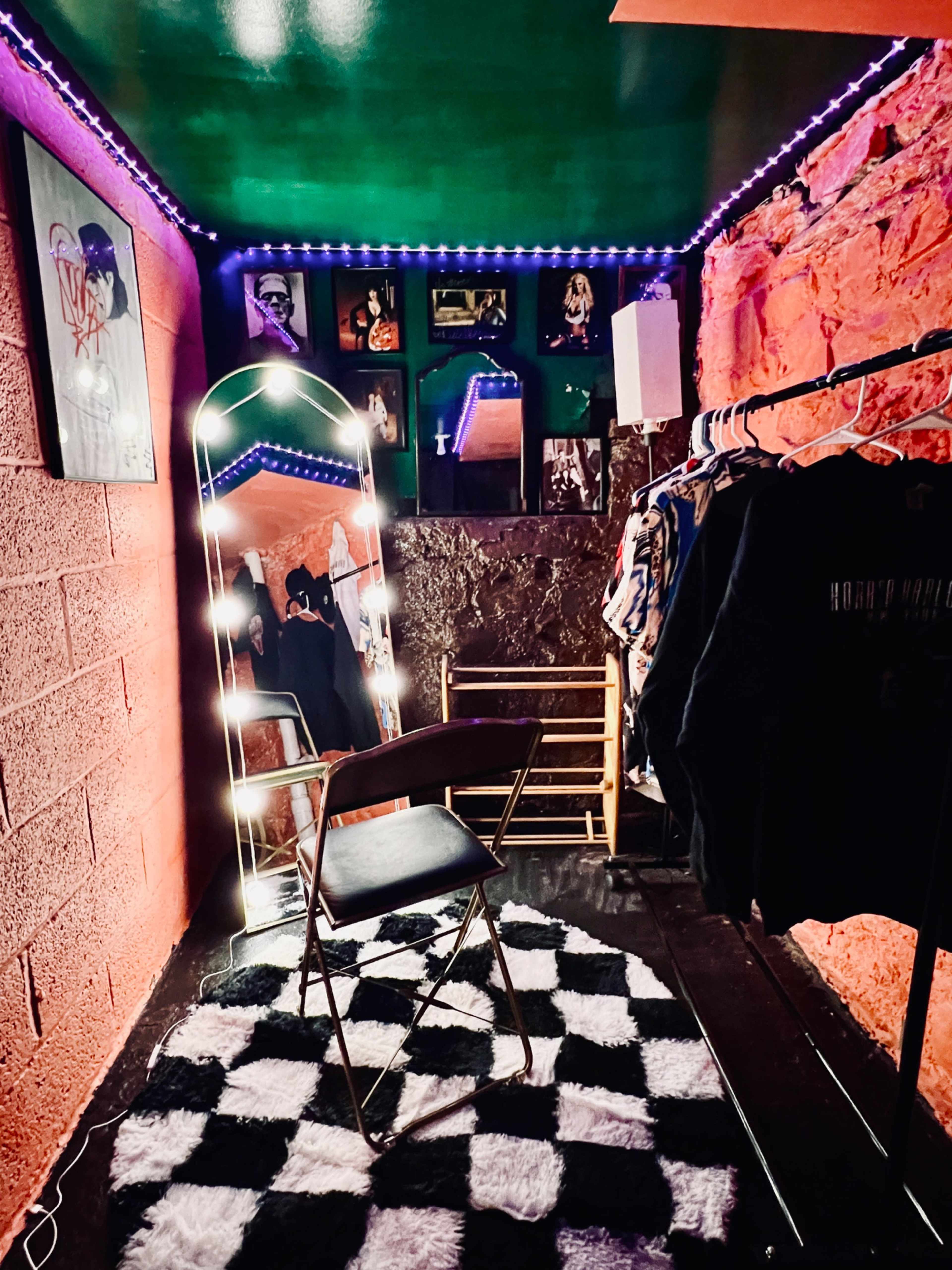 The image shows a small room with a checkered rug, a mirror framed by lights, a clothing rack with various garments, and framed artwork on the wall.