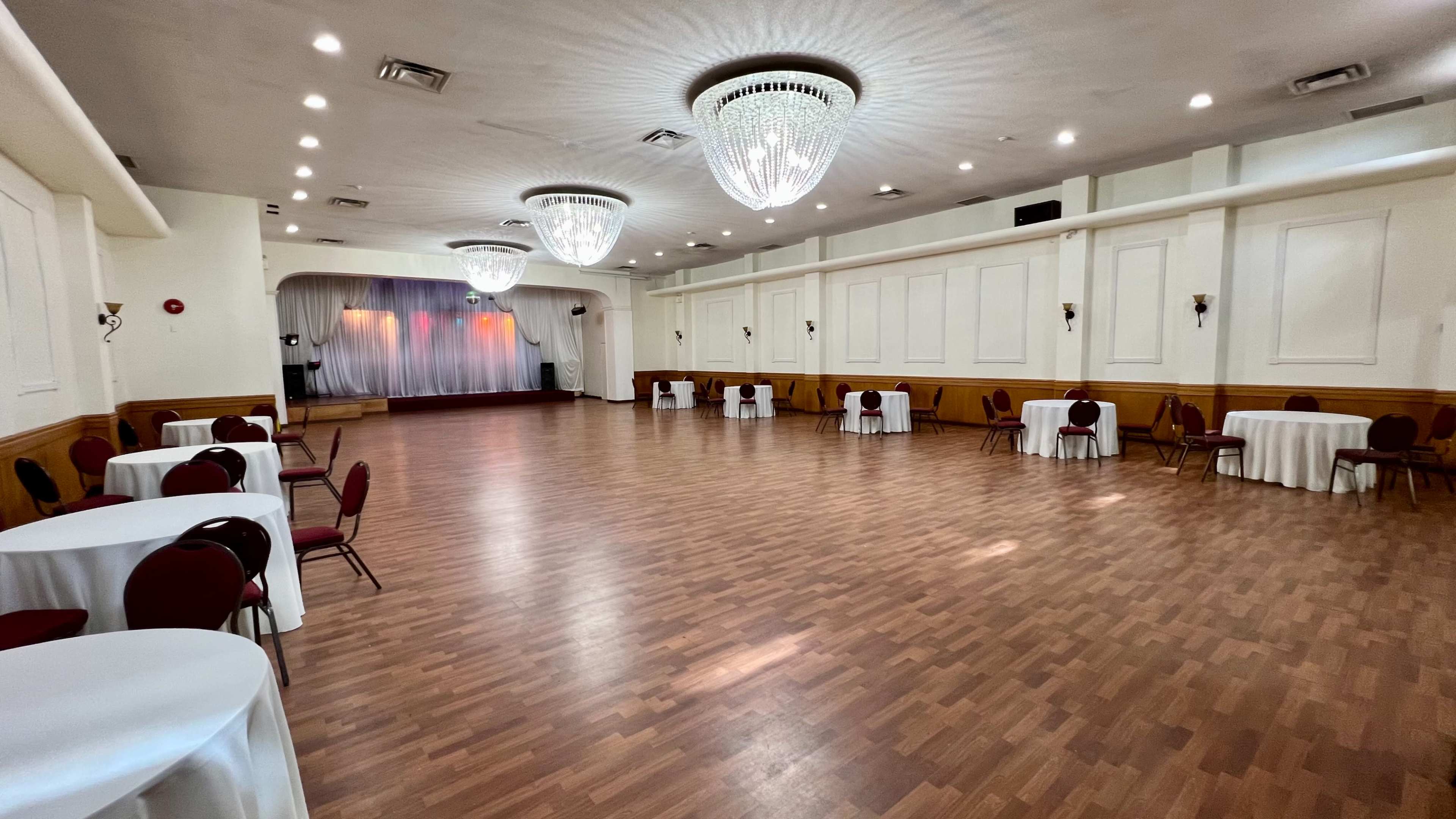 A large, empty event hall features several round tables with chairs and two chandeliers hanging from the ceiling.