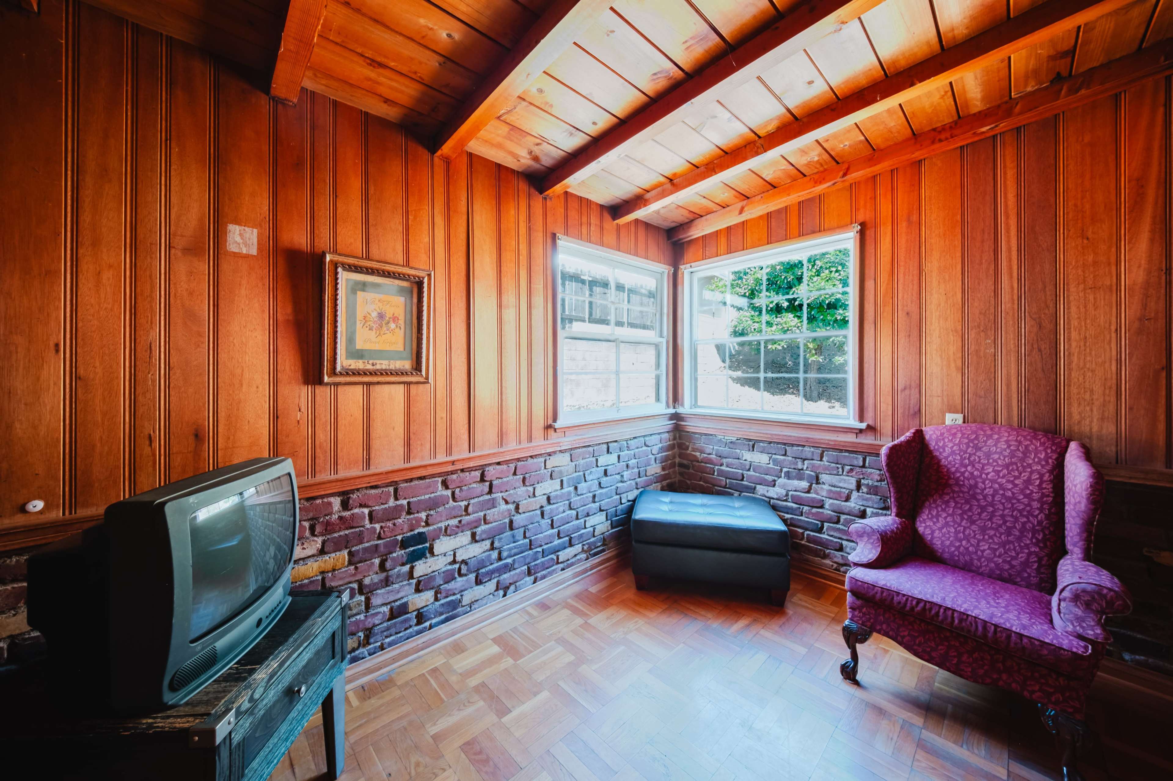 The image shows a small room with wooden walls and a sloped ceiling, featuring a vintage television, a purple armchair, and two windows allowing natural light.