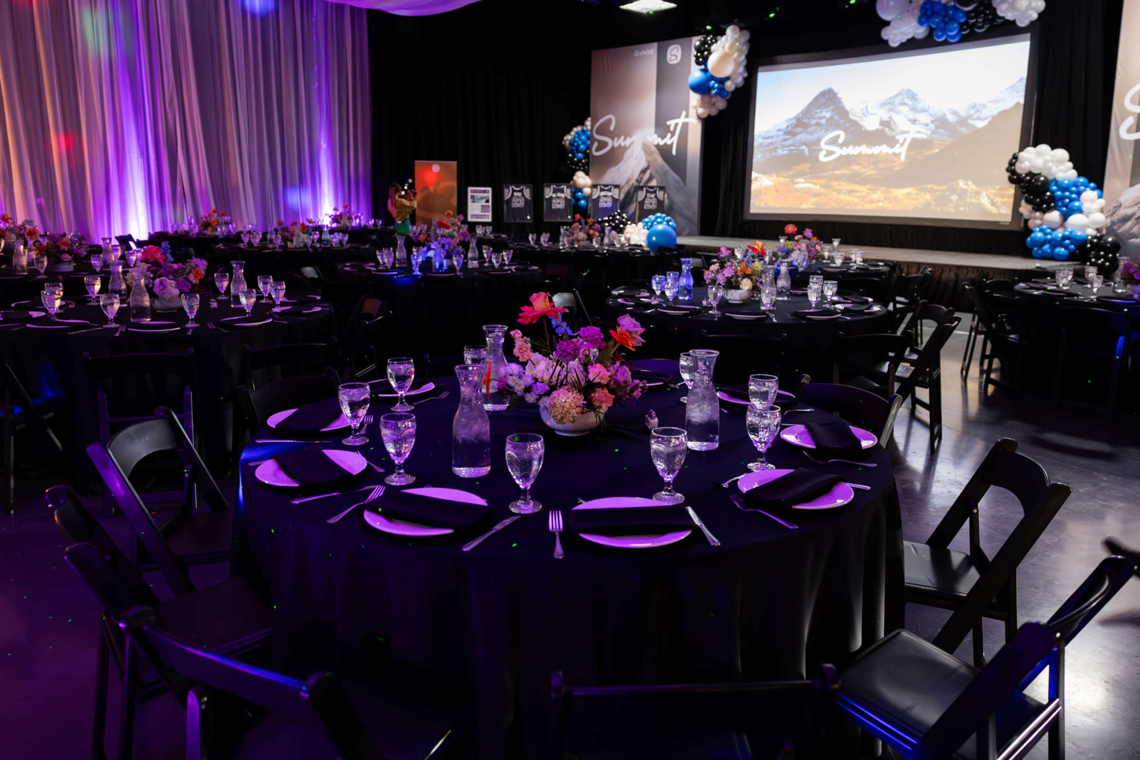 A decorated event space features round tables with black tablecloths, floral centerpieces, and glassware, set against a backdrop of screens displaying mountain imagery.