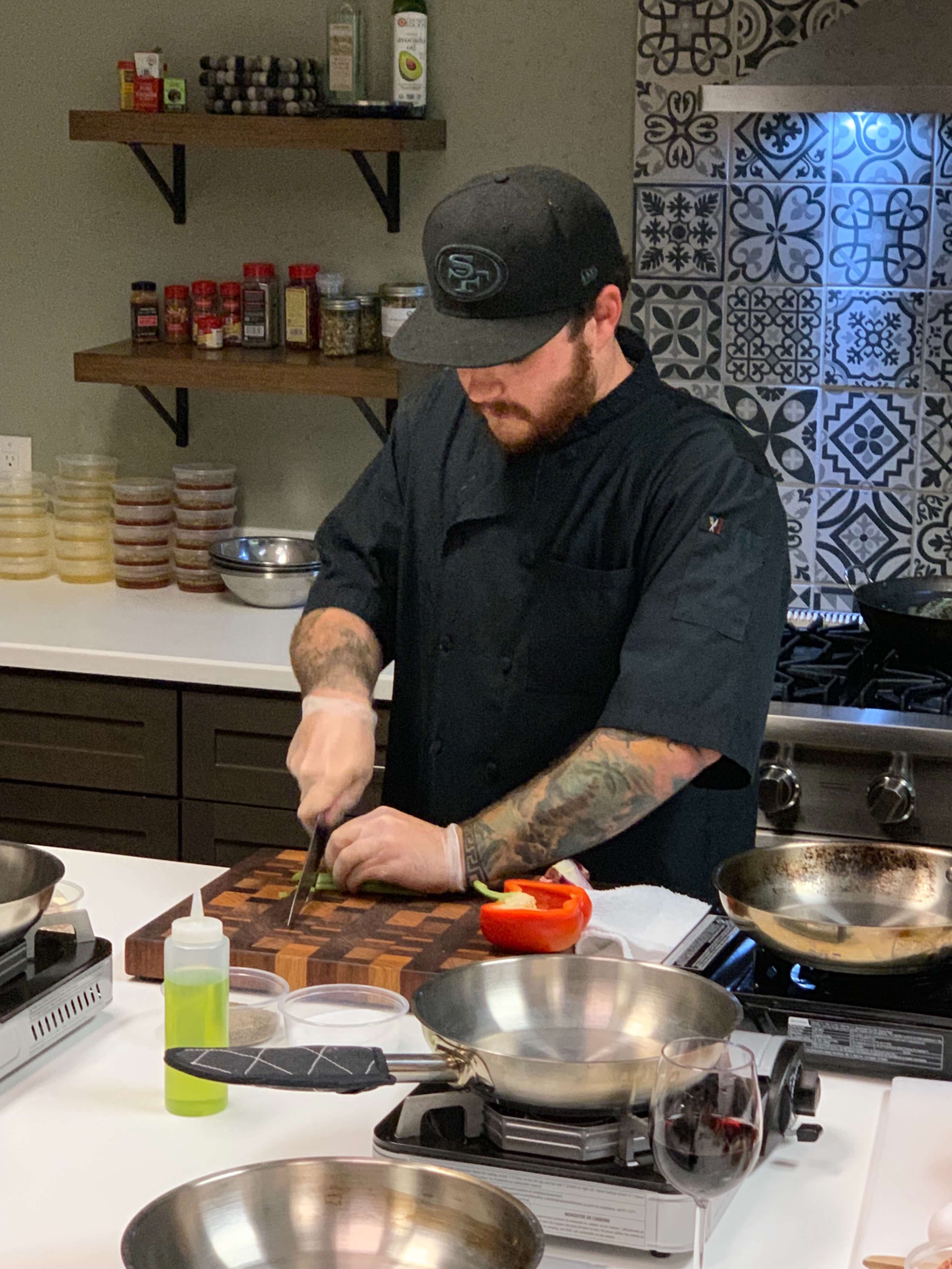A chef in a black hat prepares ingredients on a wooden cutting board in a kitchen with stainless steel appliances and patterned tiles.