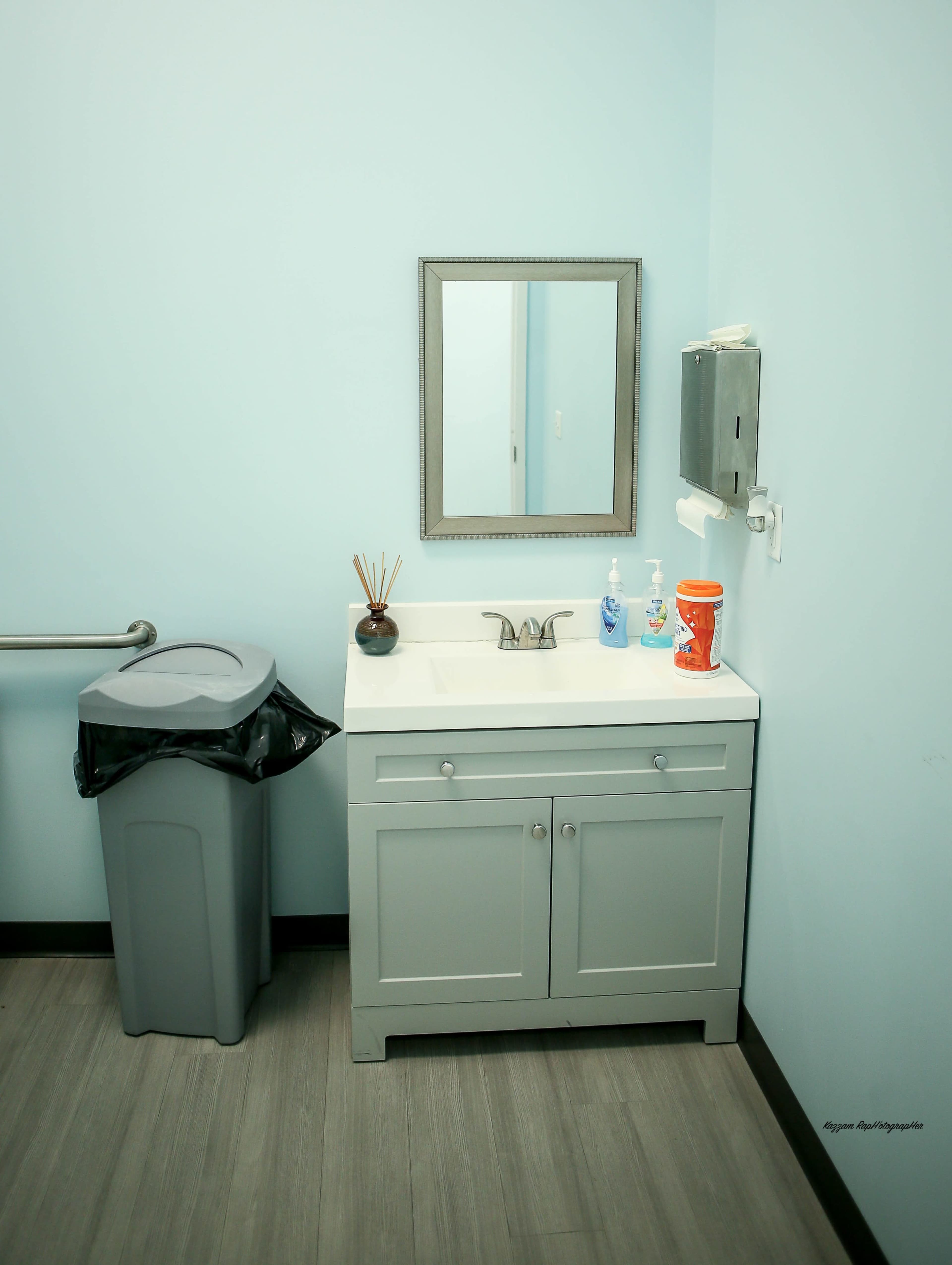 The image shows a bathroom area with a gray vanity sink, a mirror above it, a gray trash can, and a paper towel dispenser on a light blue wall.