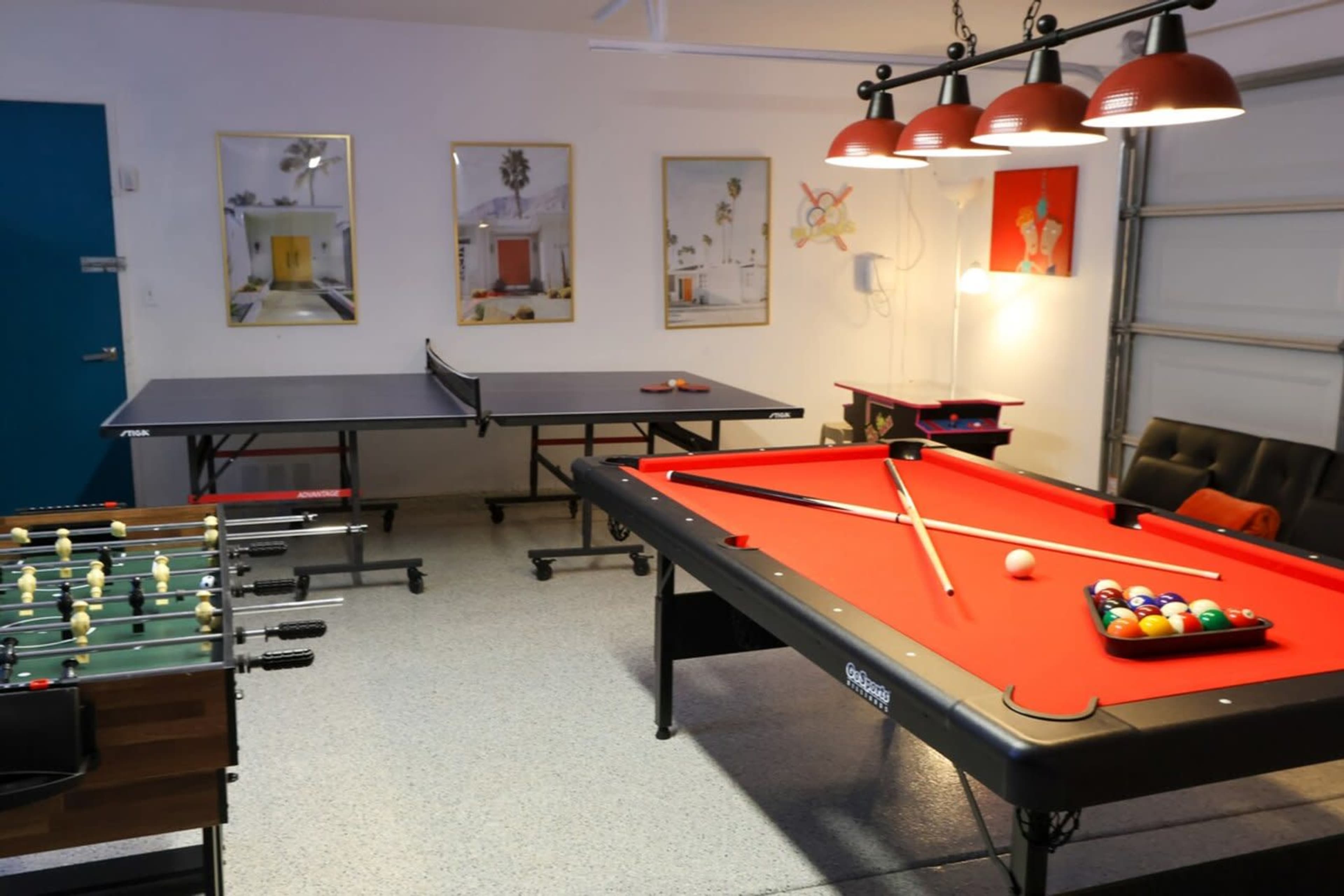 The image shows a recreational room featuring a pool table, a ping pong table, and a foosball table, along with decorative wall art and overhead lighting.