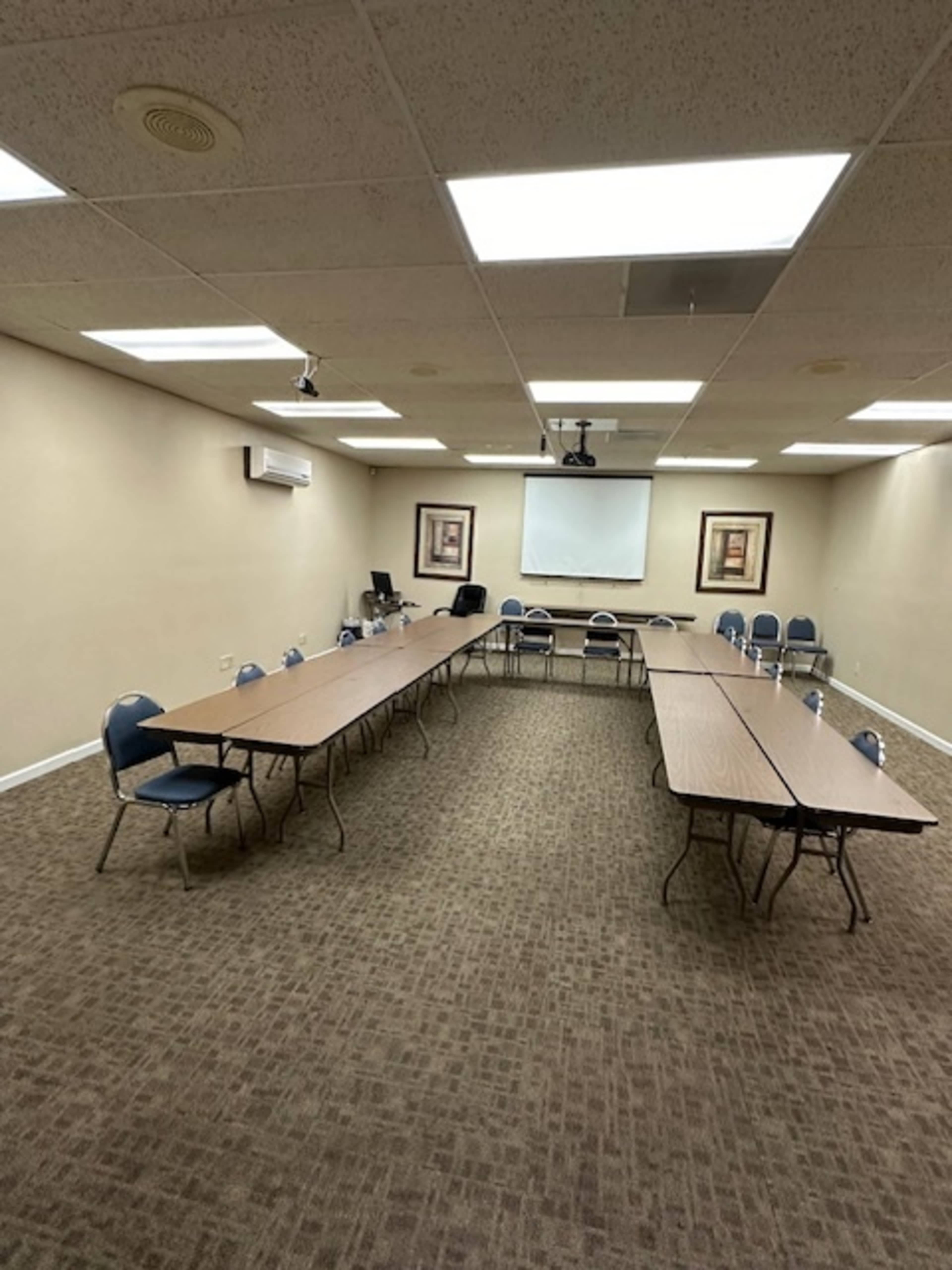 A conference room with two long tables arranged in an "H" shape, surrounded by chairs, and a projector screen at the front.