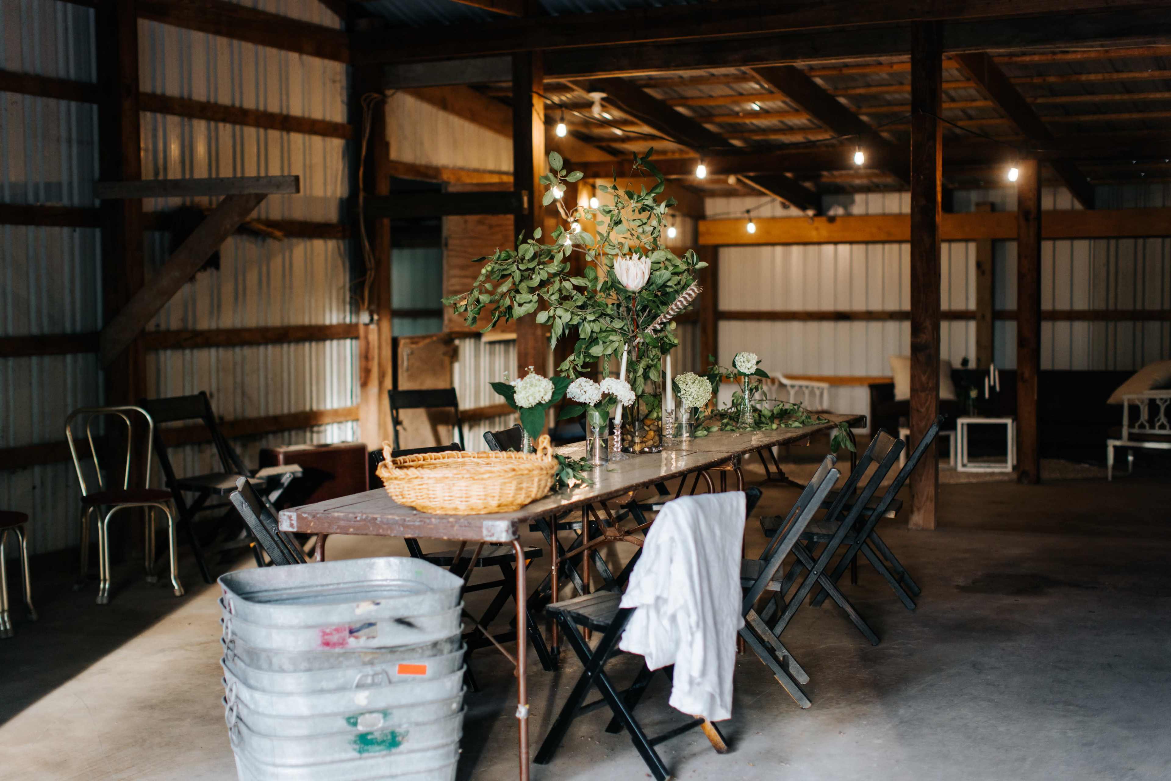 A long, wooden table is set with vases of flowers amid a rustic barn interior, featuring metal chairs and soft lighting.