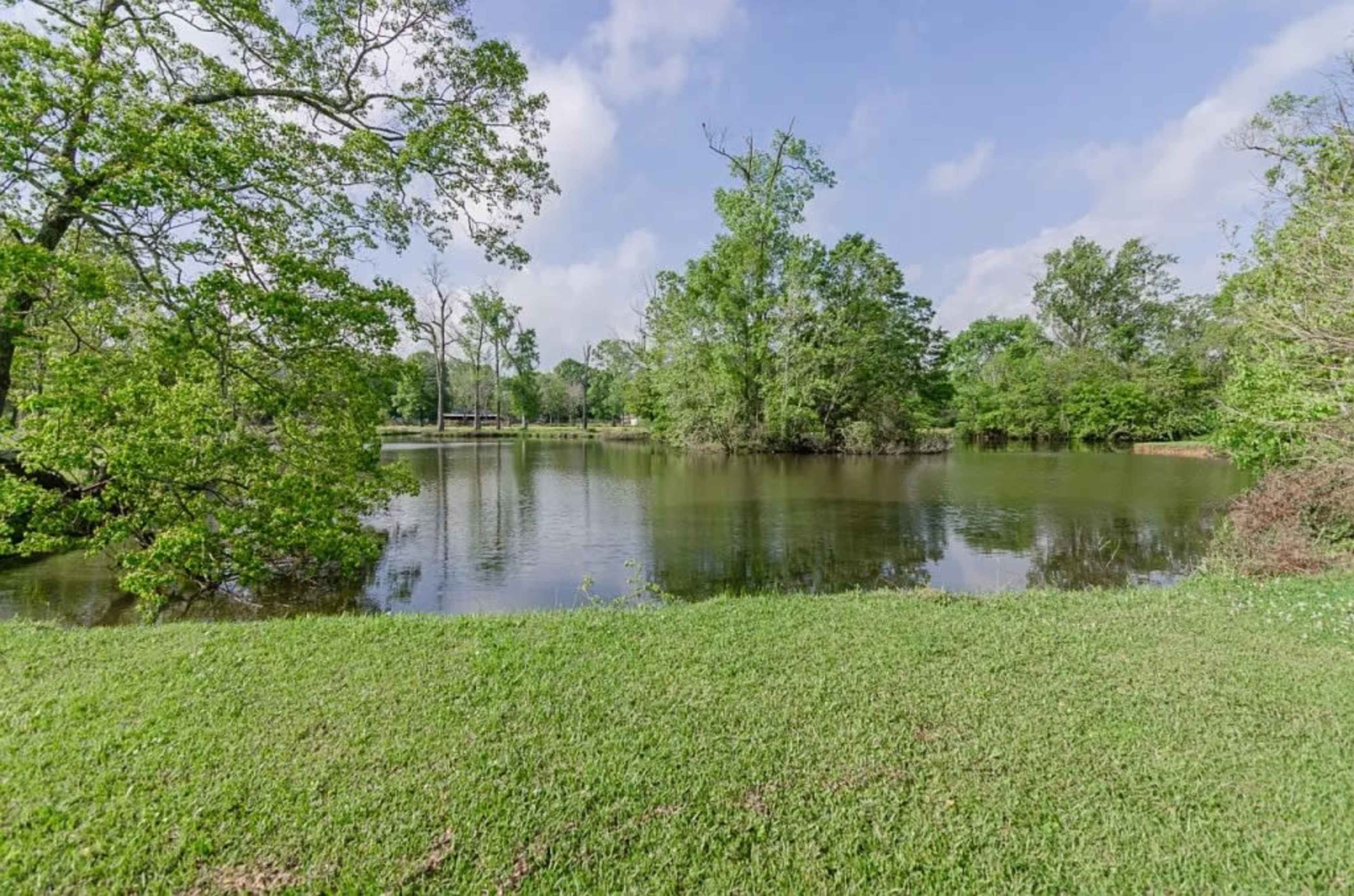 A serene lakeside scene features calm water surrounded by lush green trees and grass under a partly cloudy sky.