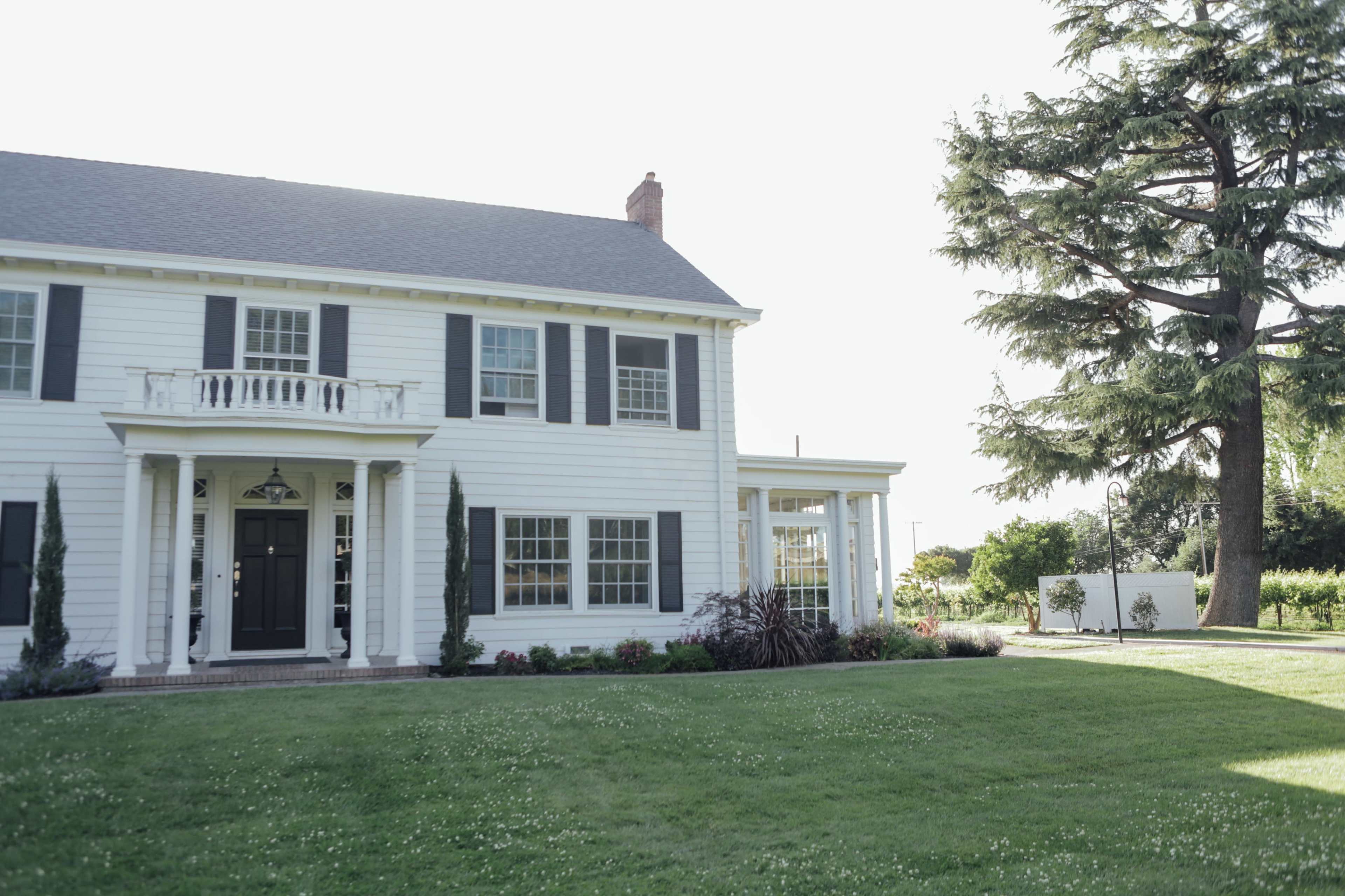 A two-story white house with dark green shutters, a front porch, and a well-manicured lawn is seen next to a large tree and garden.