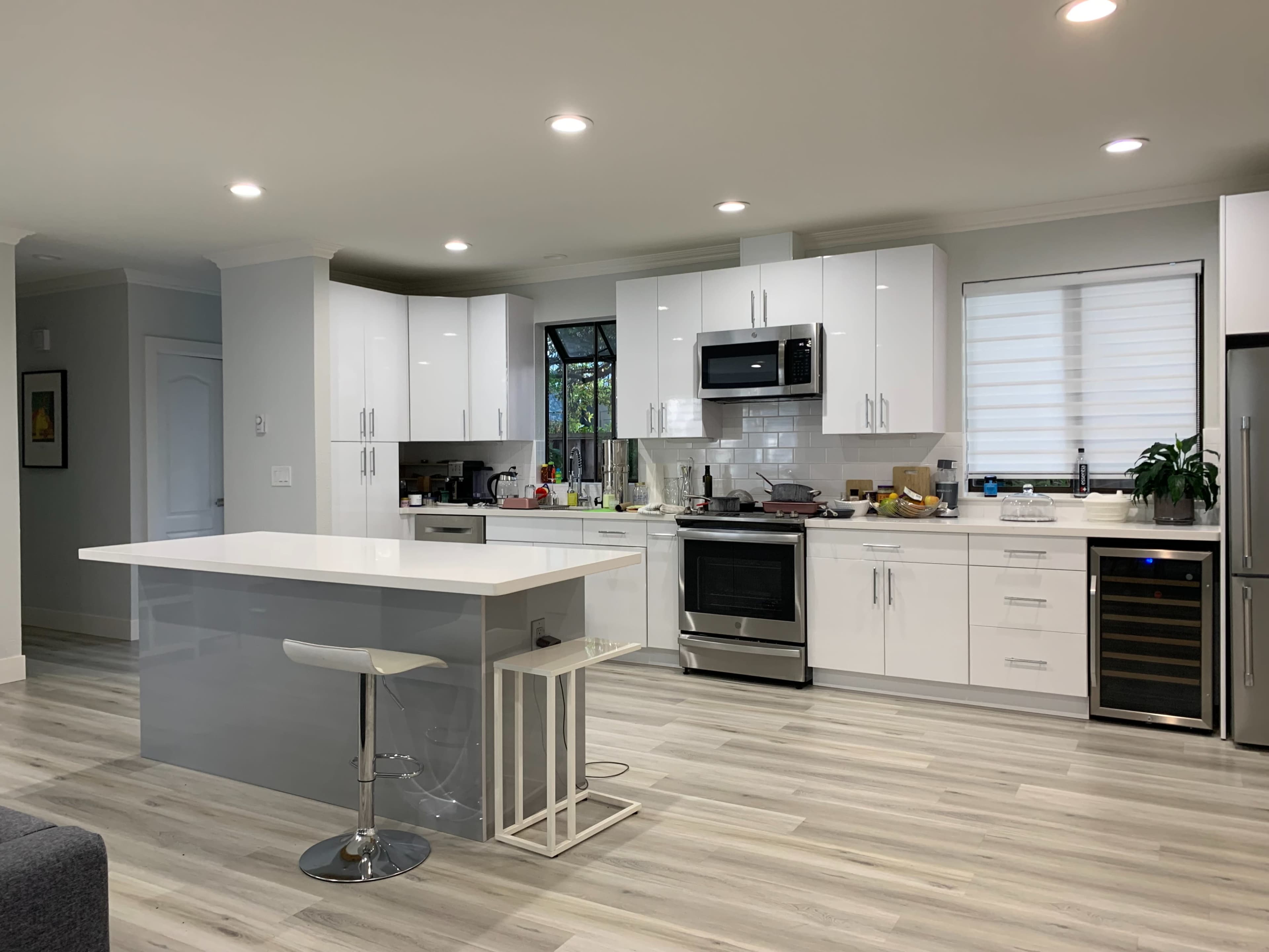A modern kitchen with white cabinets, stainless steel appliances, a central island with bar seating, and gray wood-like flooring.