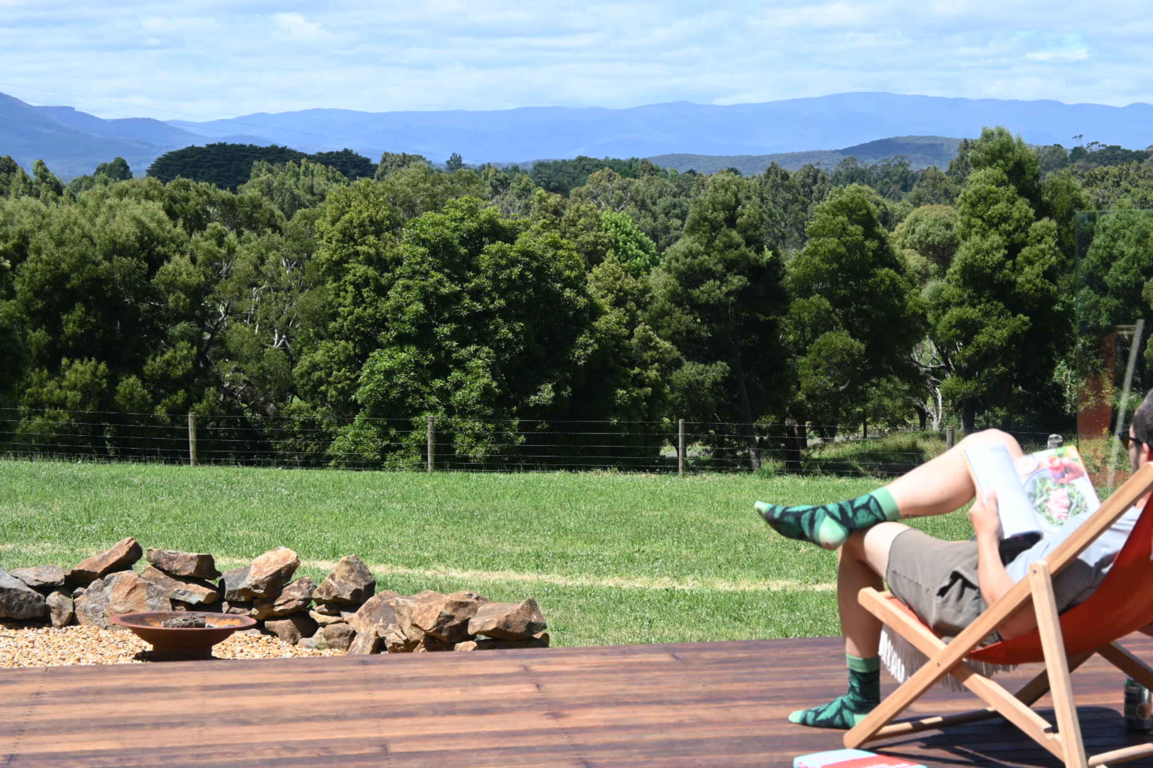 A person relaxes in a deck chair on a wooden deck overlooking a green landscape with trees and distant mountains.