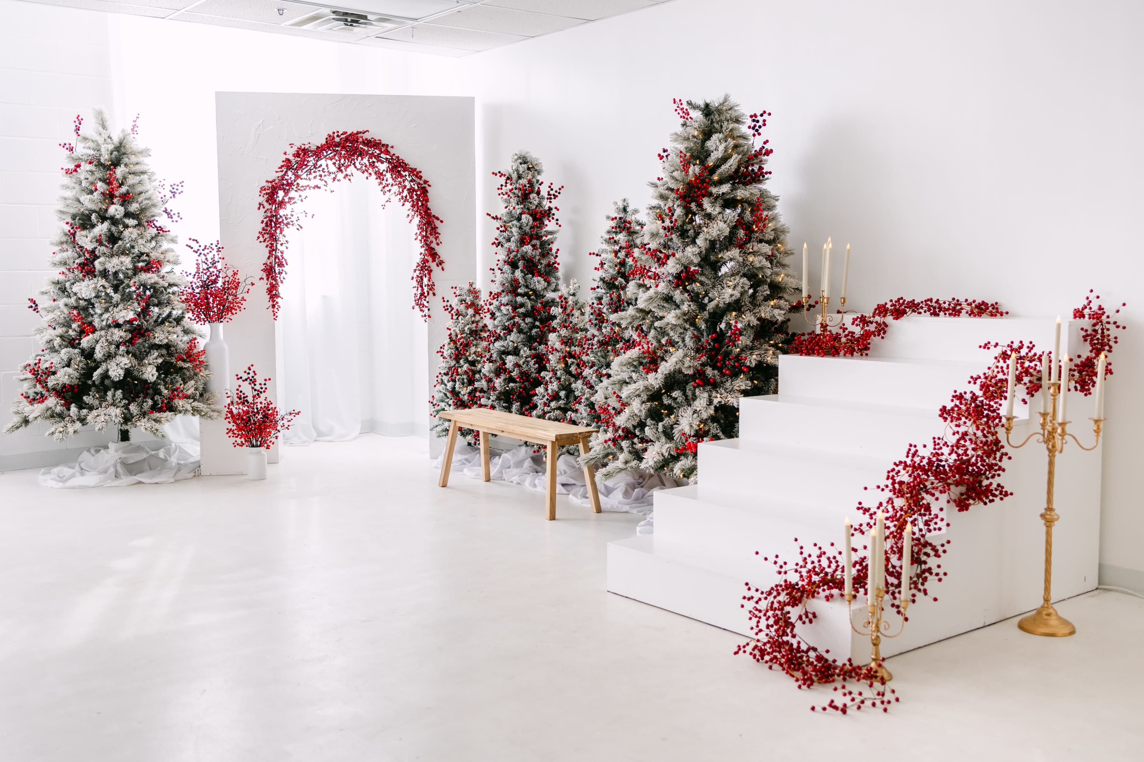 The scene features a minimalist white room decorated with snow-dusted Christmas trees, red floral arrangements, a staircase, a wooden bench, and golden candle holders.