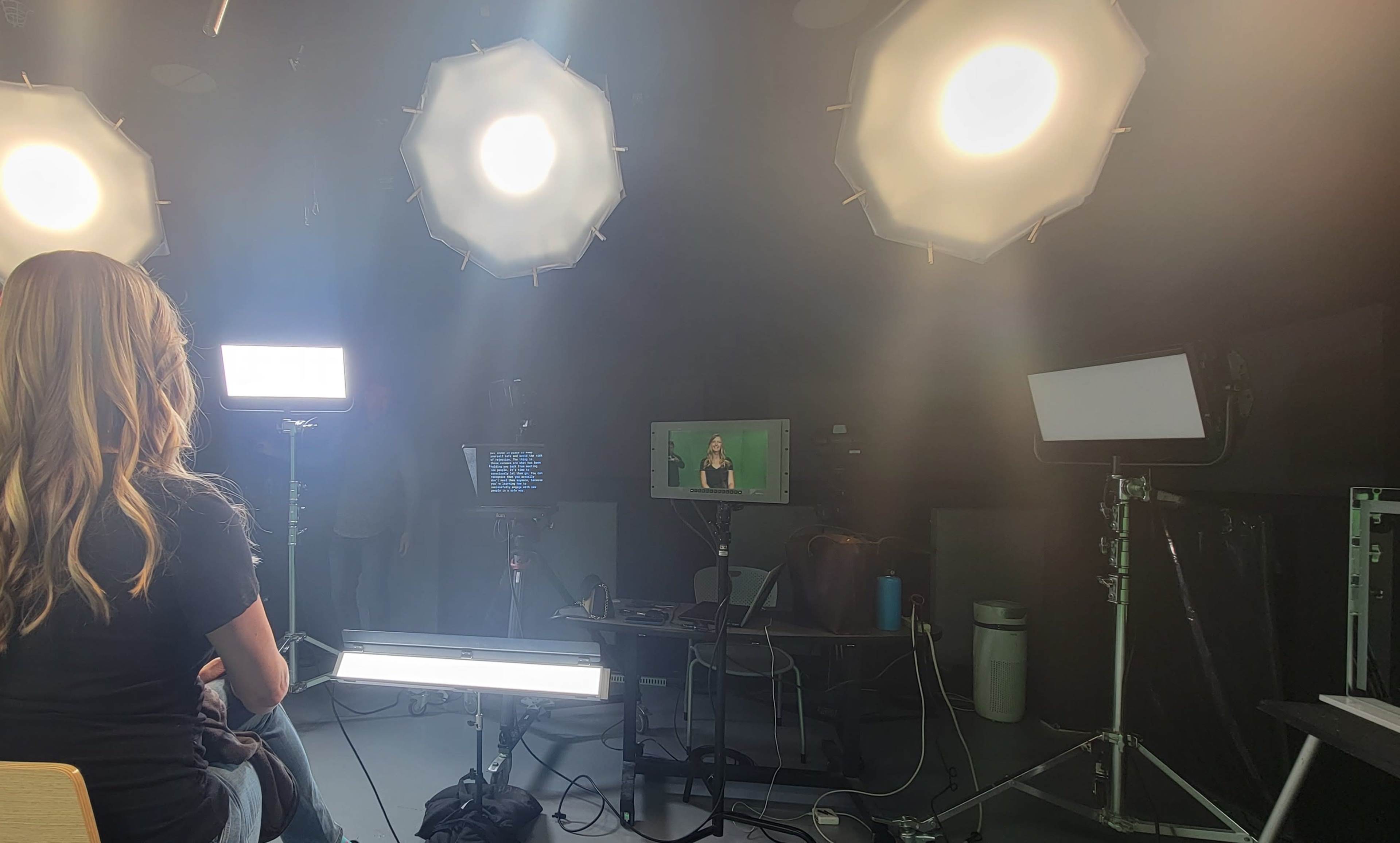 A person sits in a studio illuminated by multiple softbox lights, with a video monitor displaying a speaker on a green screen backdrop.