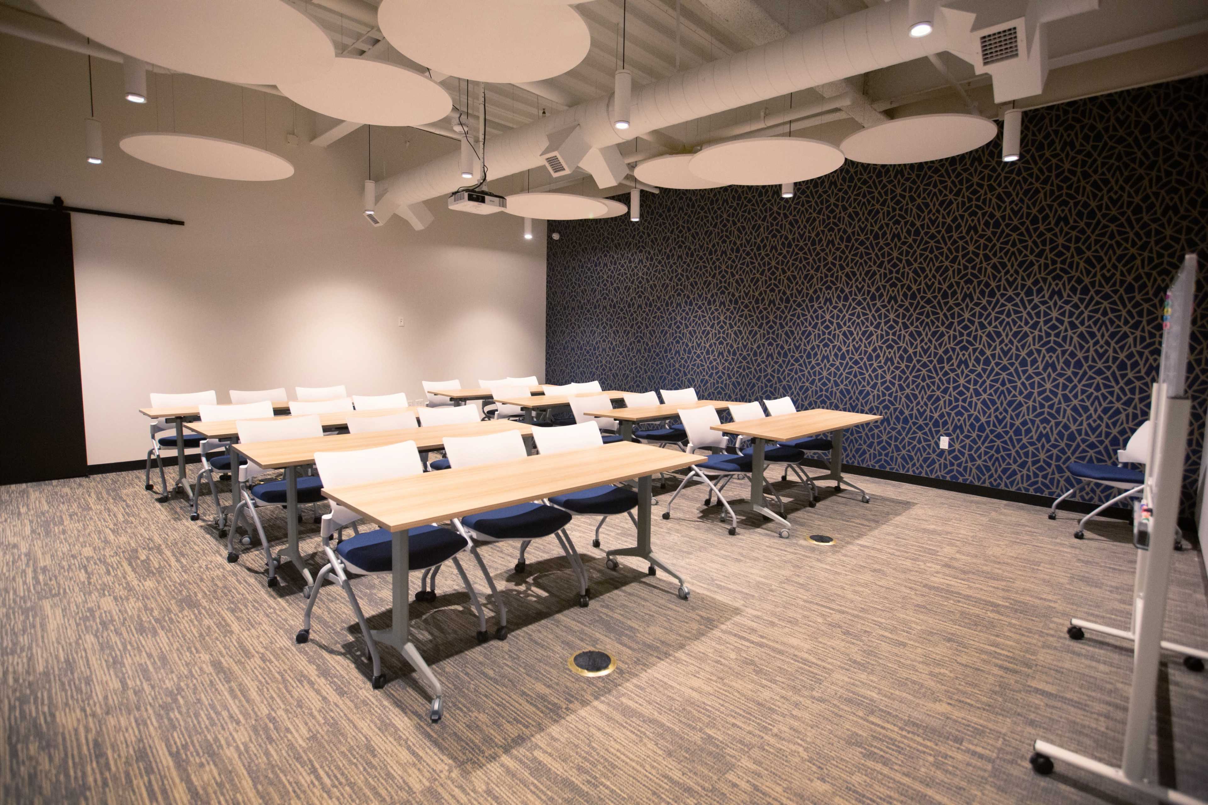 A classroom features several rows of tables and chairs, with a textured blue wall in the background and acoustic ceiling panels overhead.