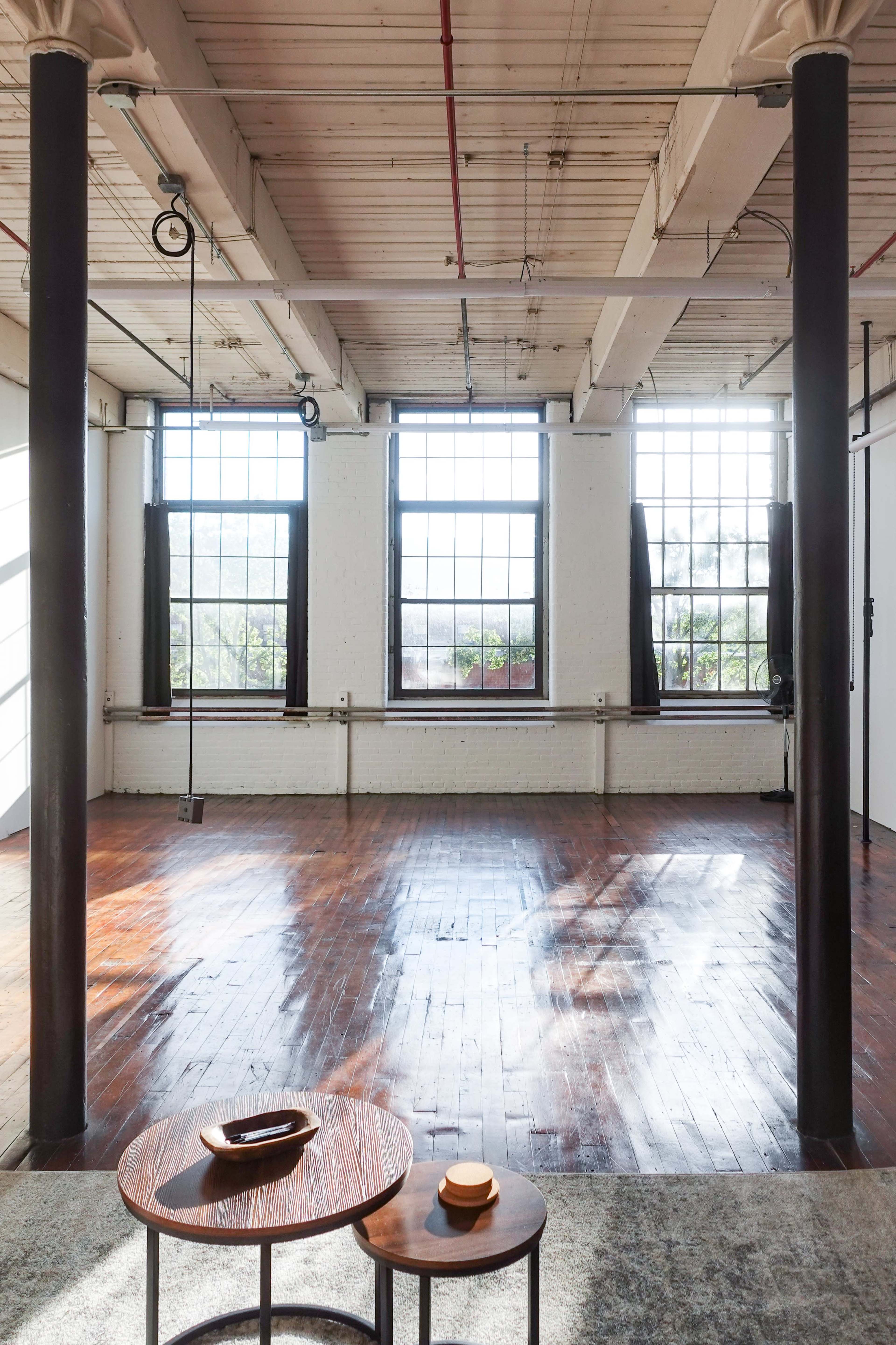 An empty industrial-style room with large windows, wooden floors, and a pair of small tables.