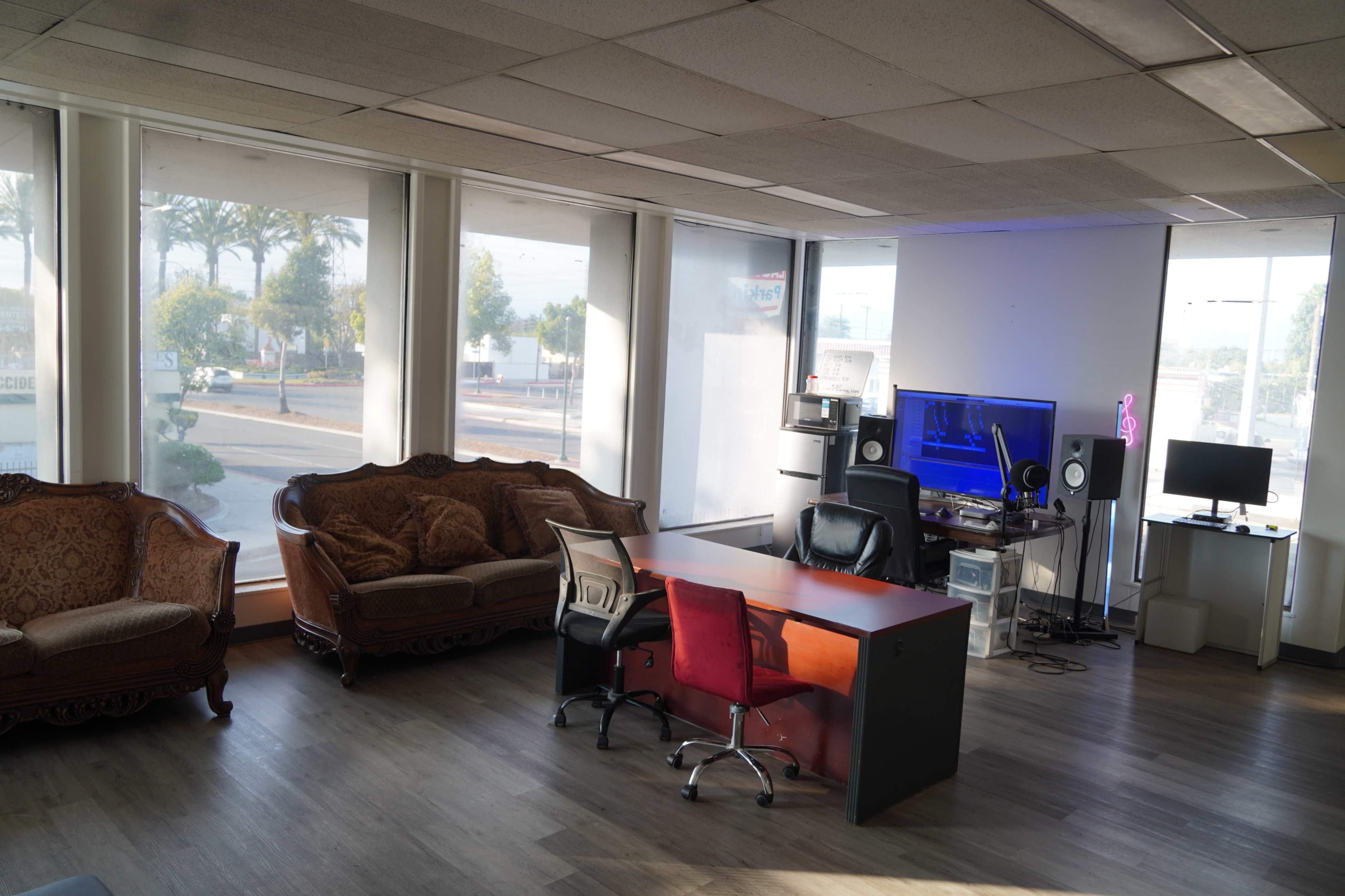 The image shows a modern office space with a large window, two sofas, a desk with chairs, and computers set up against the walls.