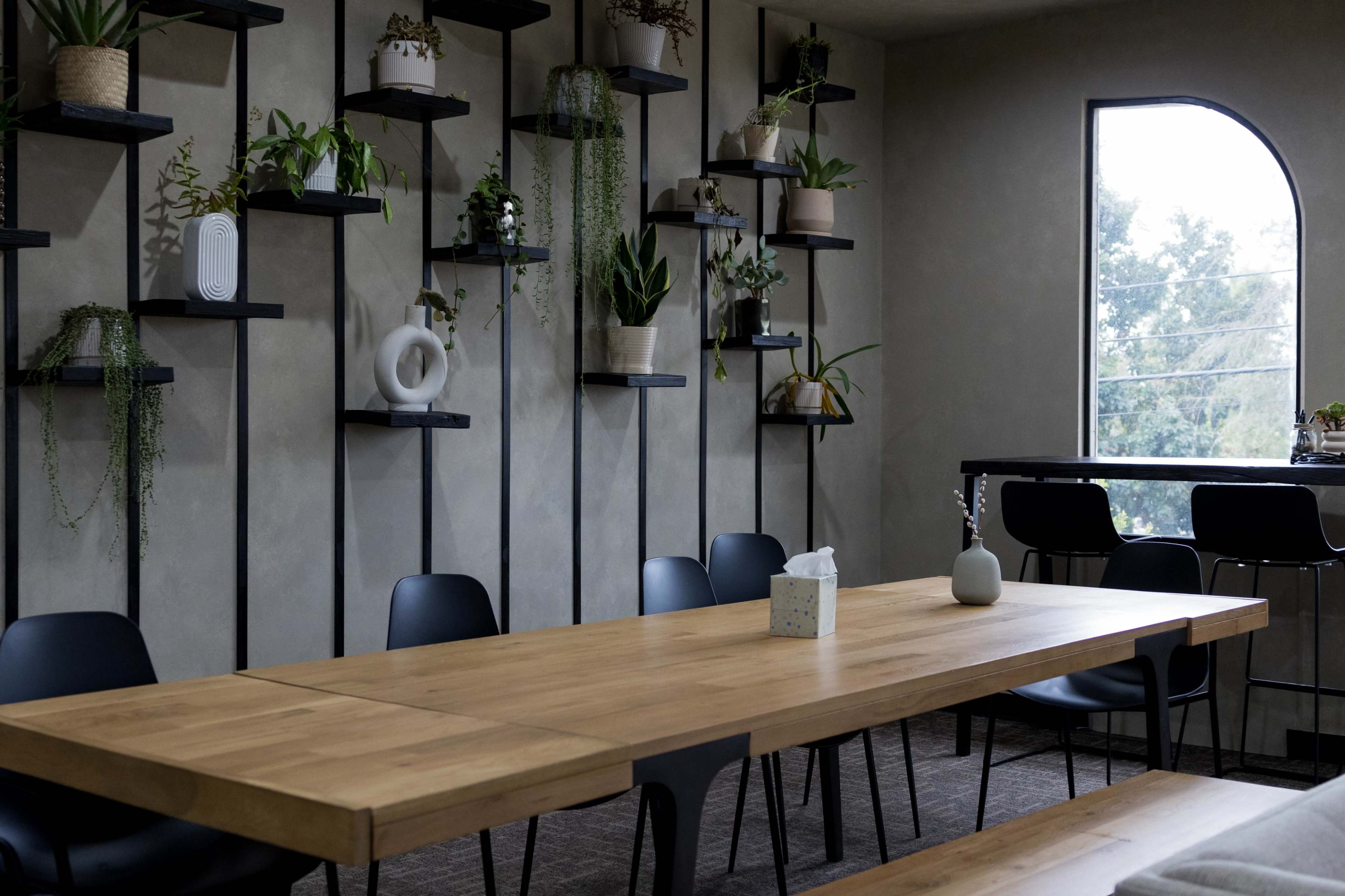 A long wooden table is set in a modern room with plants displaying on wall-mounted shelves and a bar area in the background.