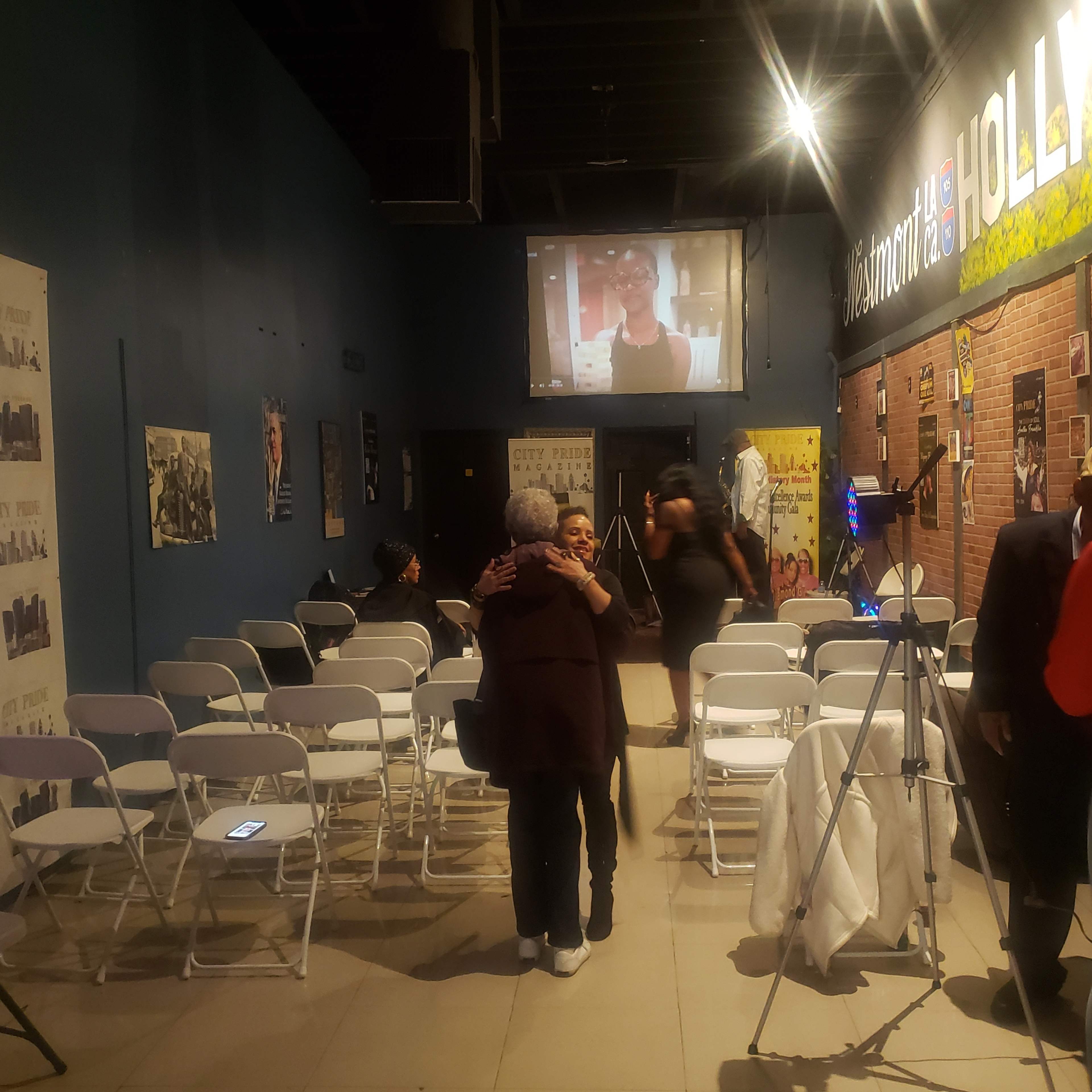 A group of people is gathered in a room with empty chairs, while one woman embraces another as a video plays on a screen in the background.