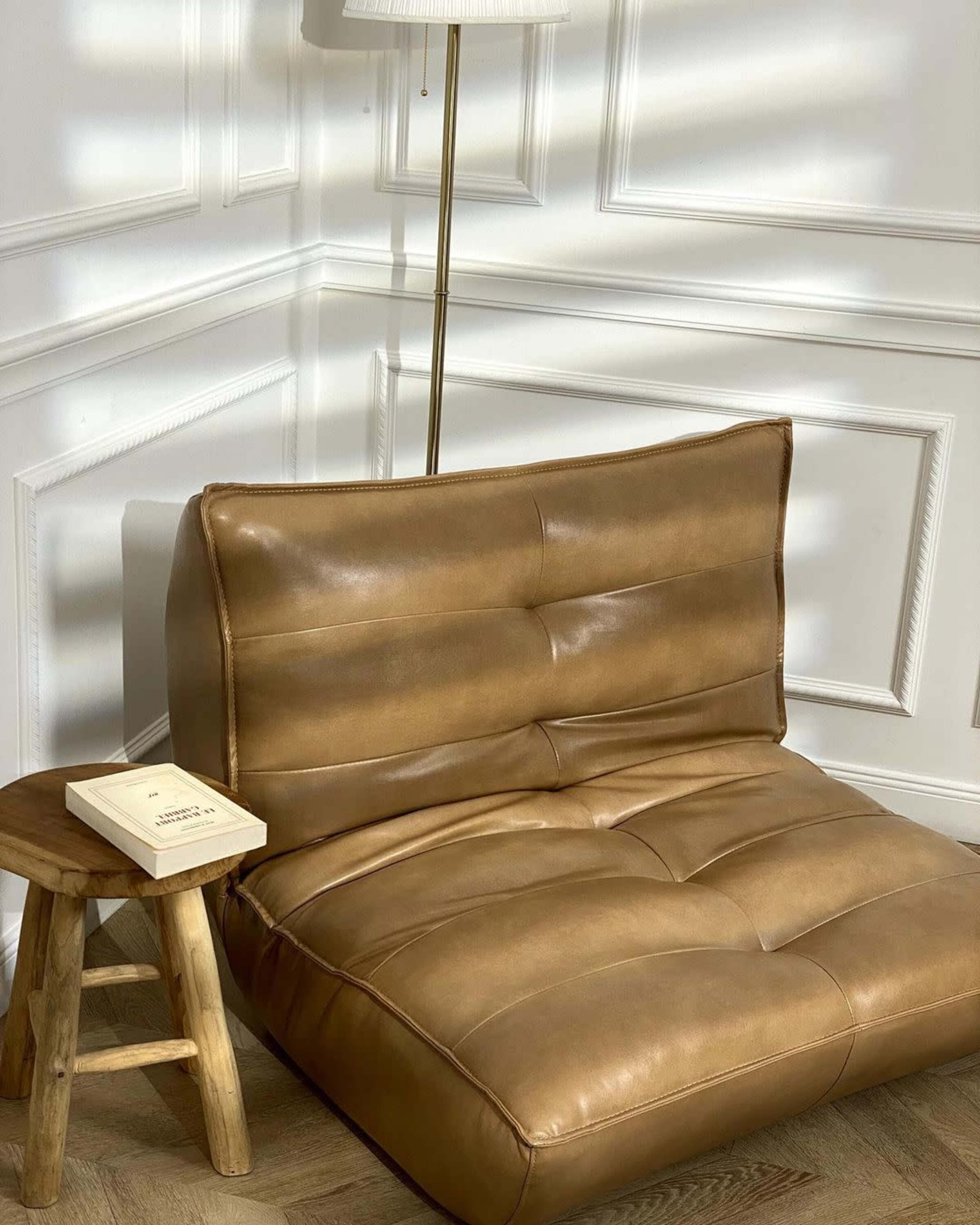 A brown leather chair is positioned next to a small wooden stool and a standing lamp in a well-lit room with paneled walls.