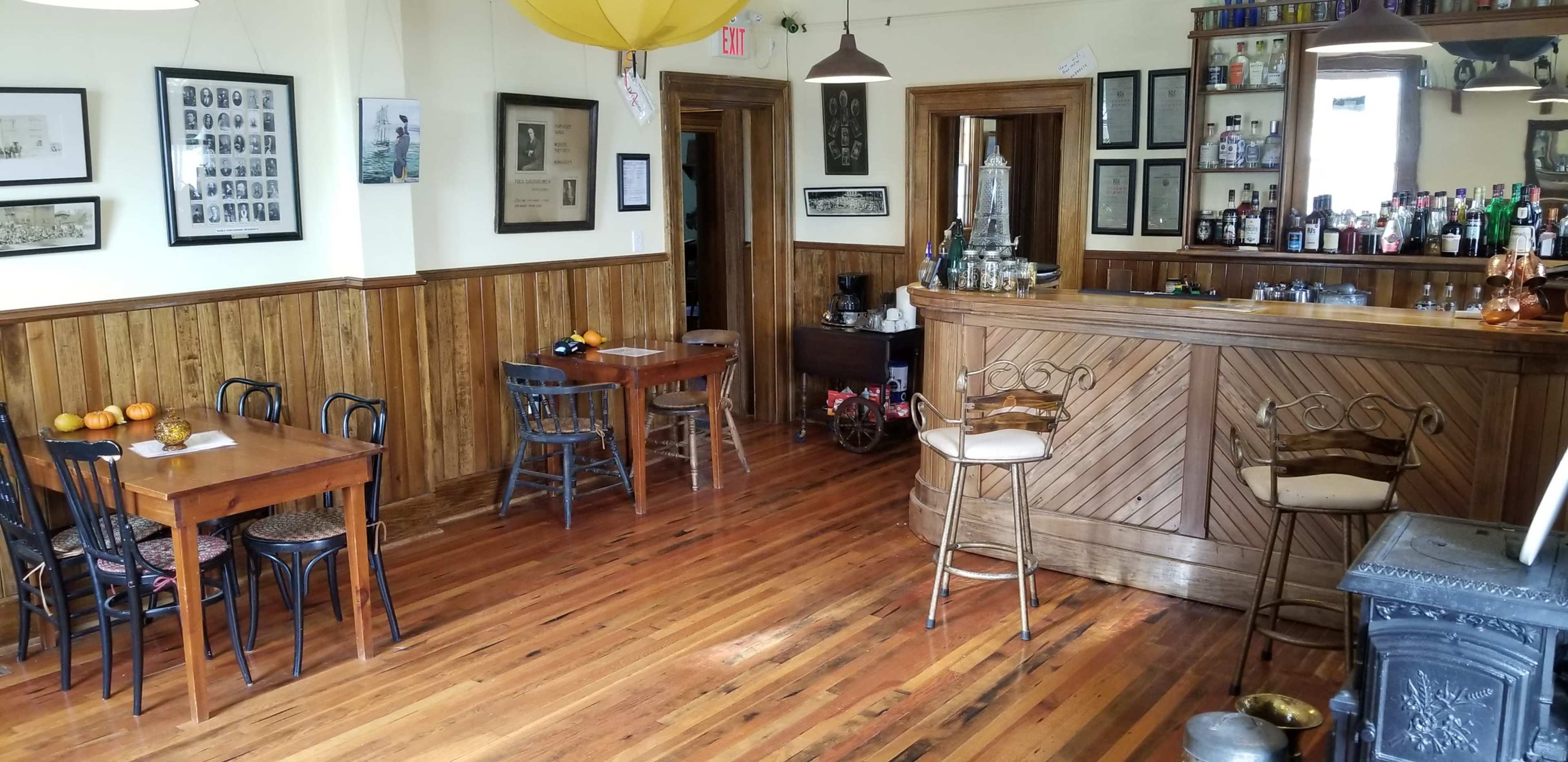 A rustic bar area with wooden flooring, several tables, and a counter stocked with various bottles.