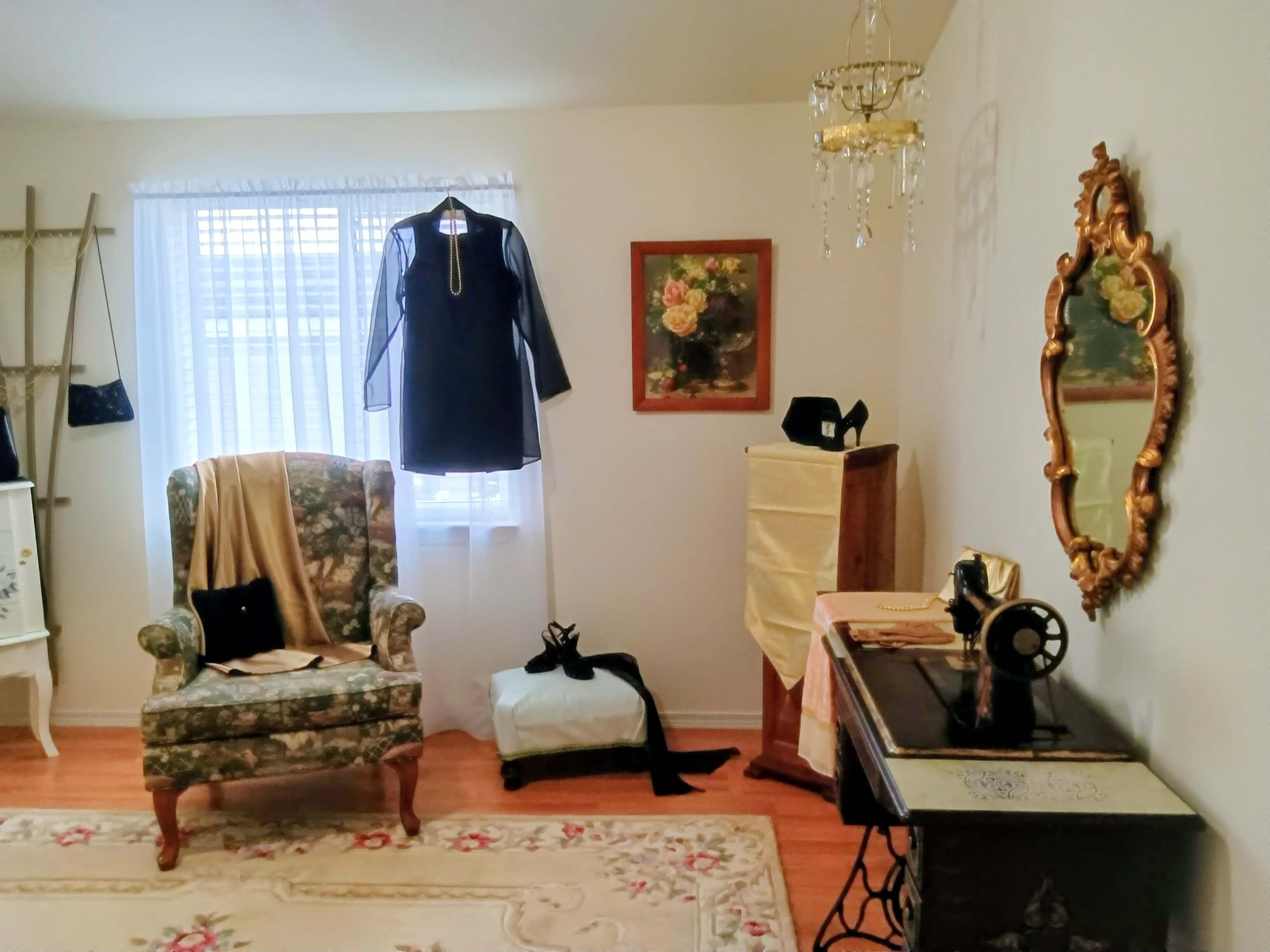 The image shows a vintage-style room with a floral armchair, a dress hanging in front of a window, and a sewing machine positioned beside a wooden cabinet.