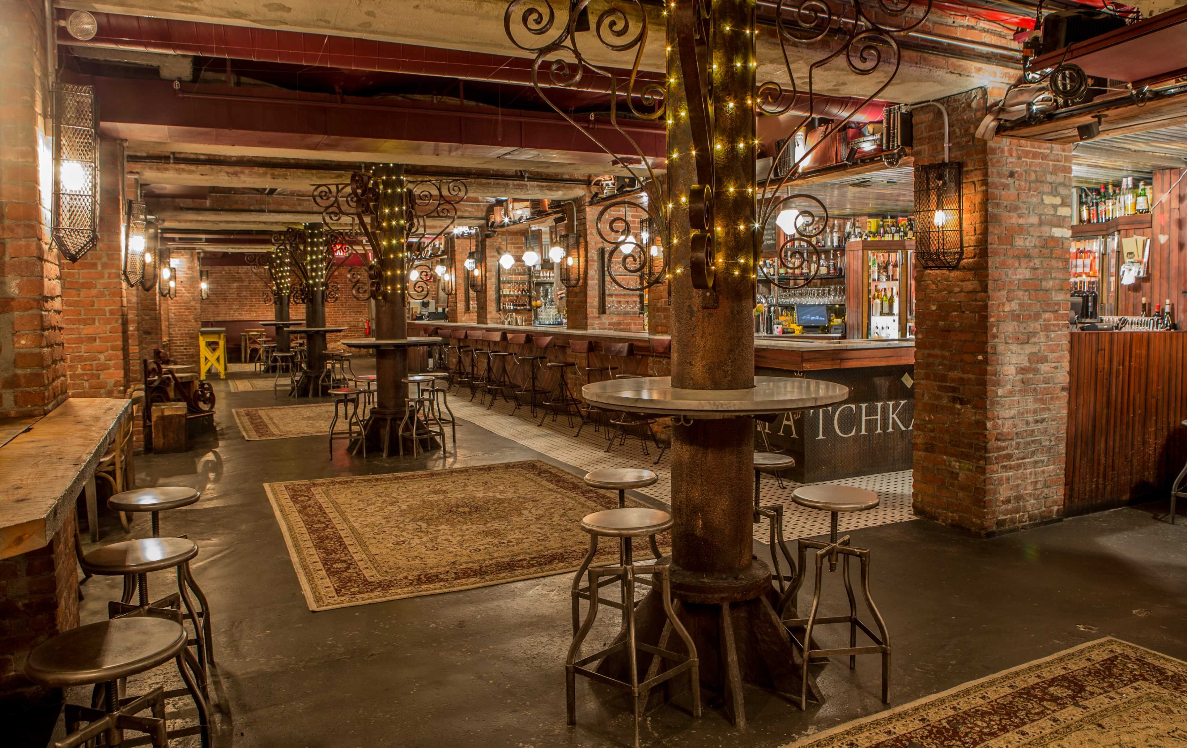 The image shows a dimly lit bar interior with exposed brick walls, wooden tables, and metal stools arranged around decorative columns illuminated by string lights.