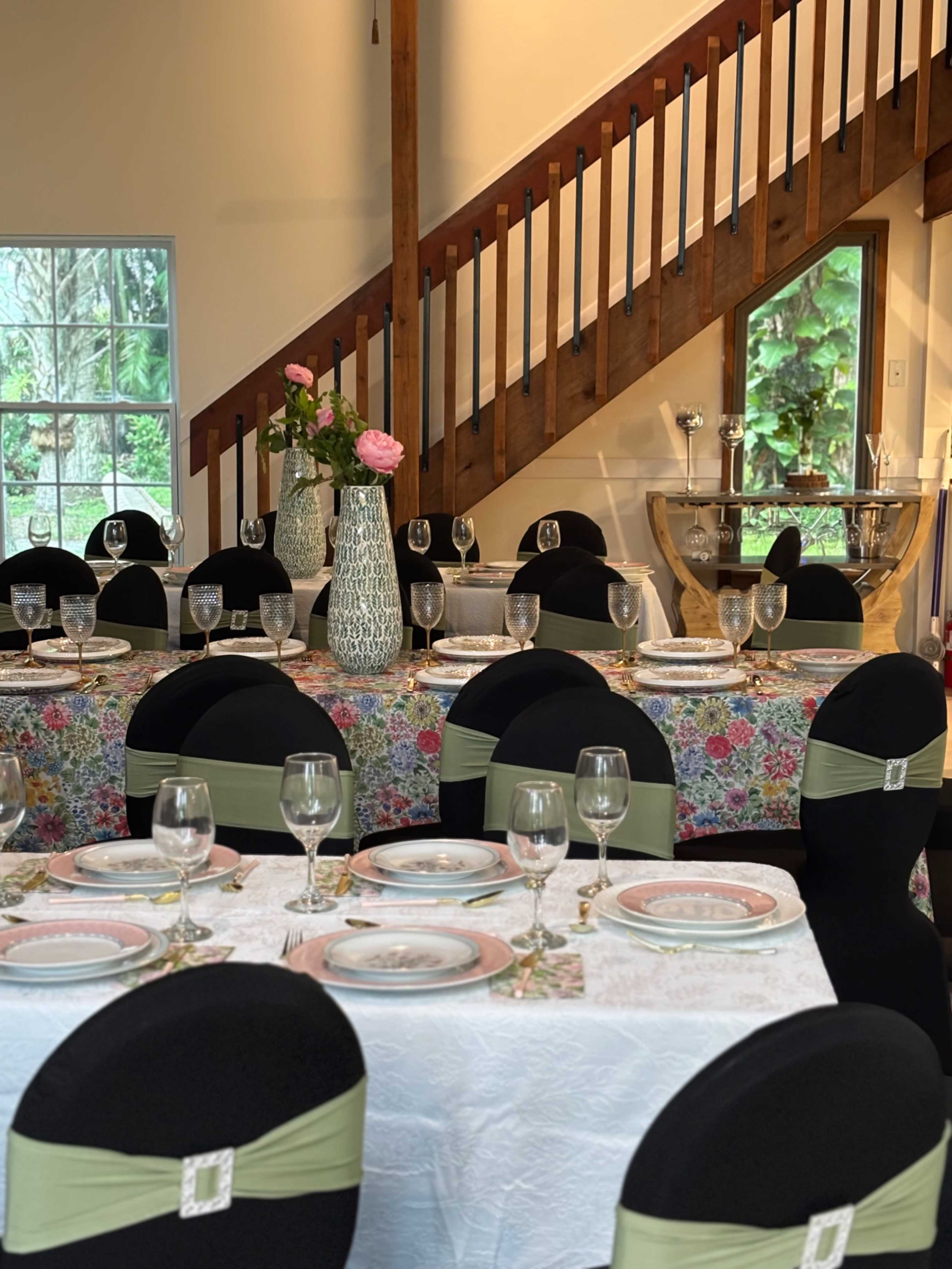The image shows a dining area with elegantly set tables adorned with floral tablecloths, wine glasses, and decorative centerpieces, accompanied by a staircase in the background.