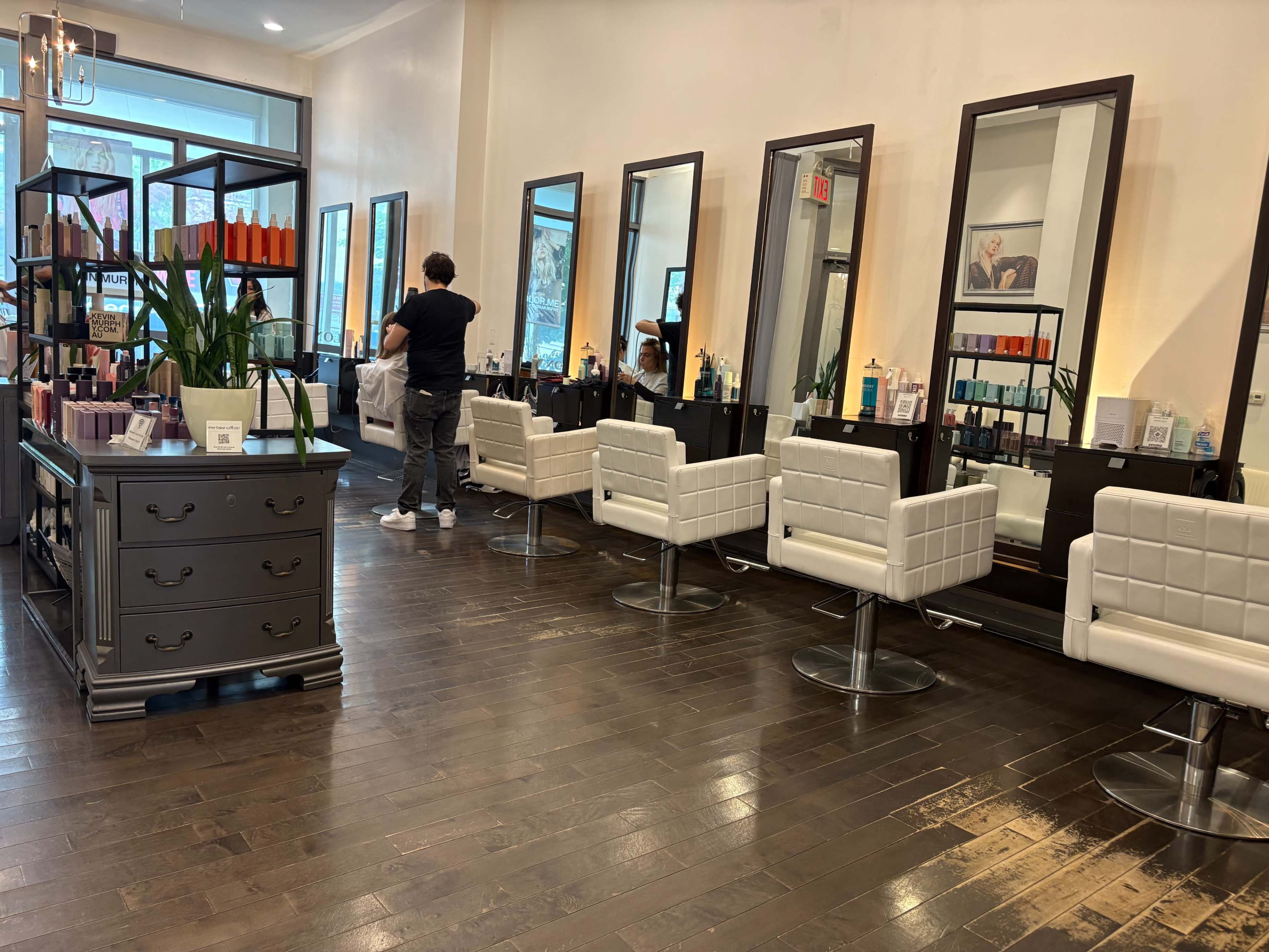 The image shows a modern hair salon with white chairs, large mirrors, and shelves displaying hair care products.