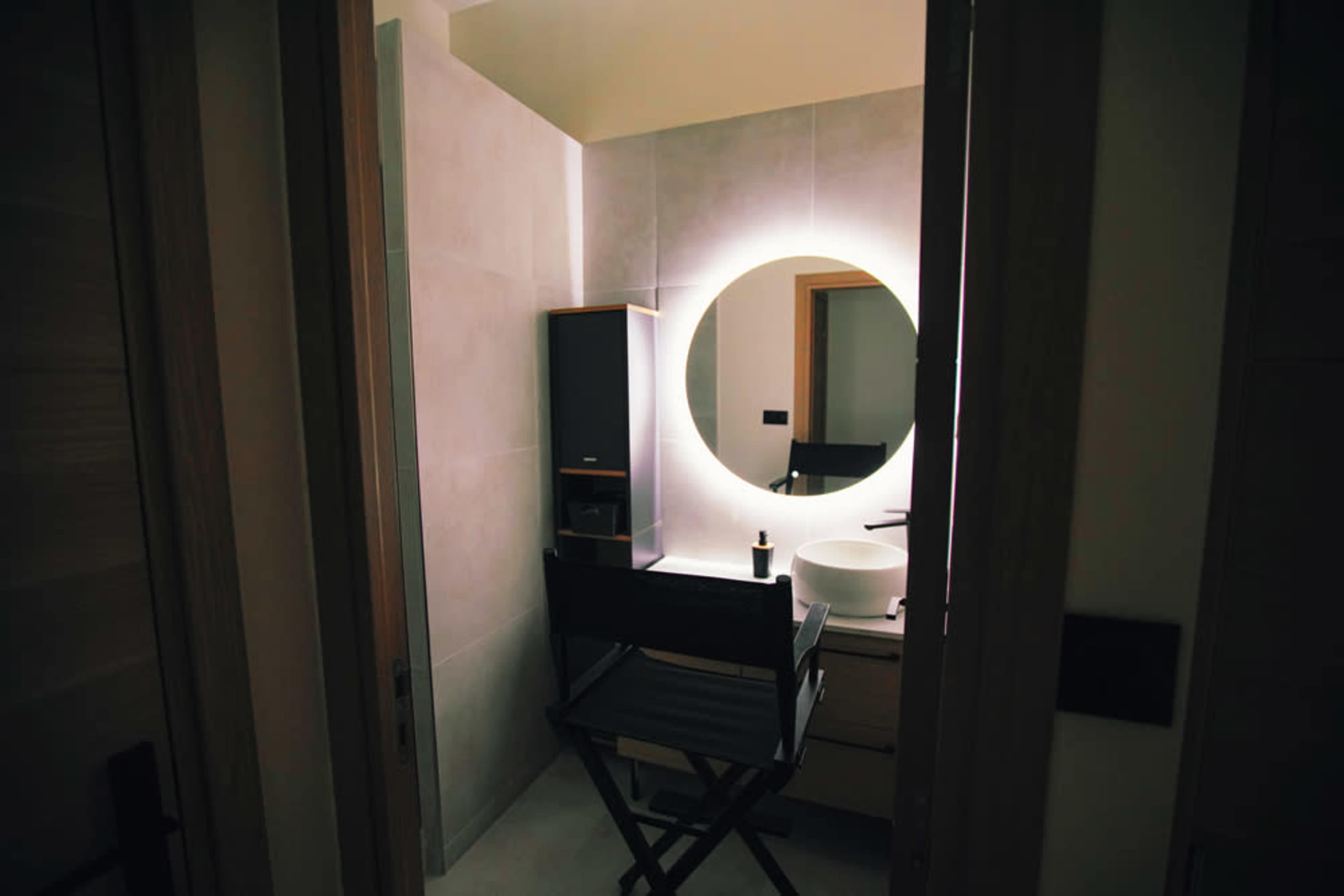 A modern bathroom features a circular illuminated mirror above a sink, with a folding chair positioned nearby.