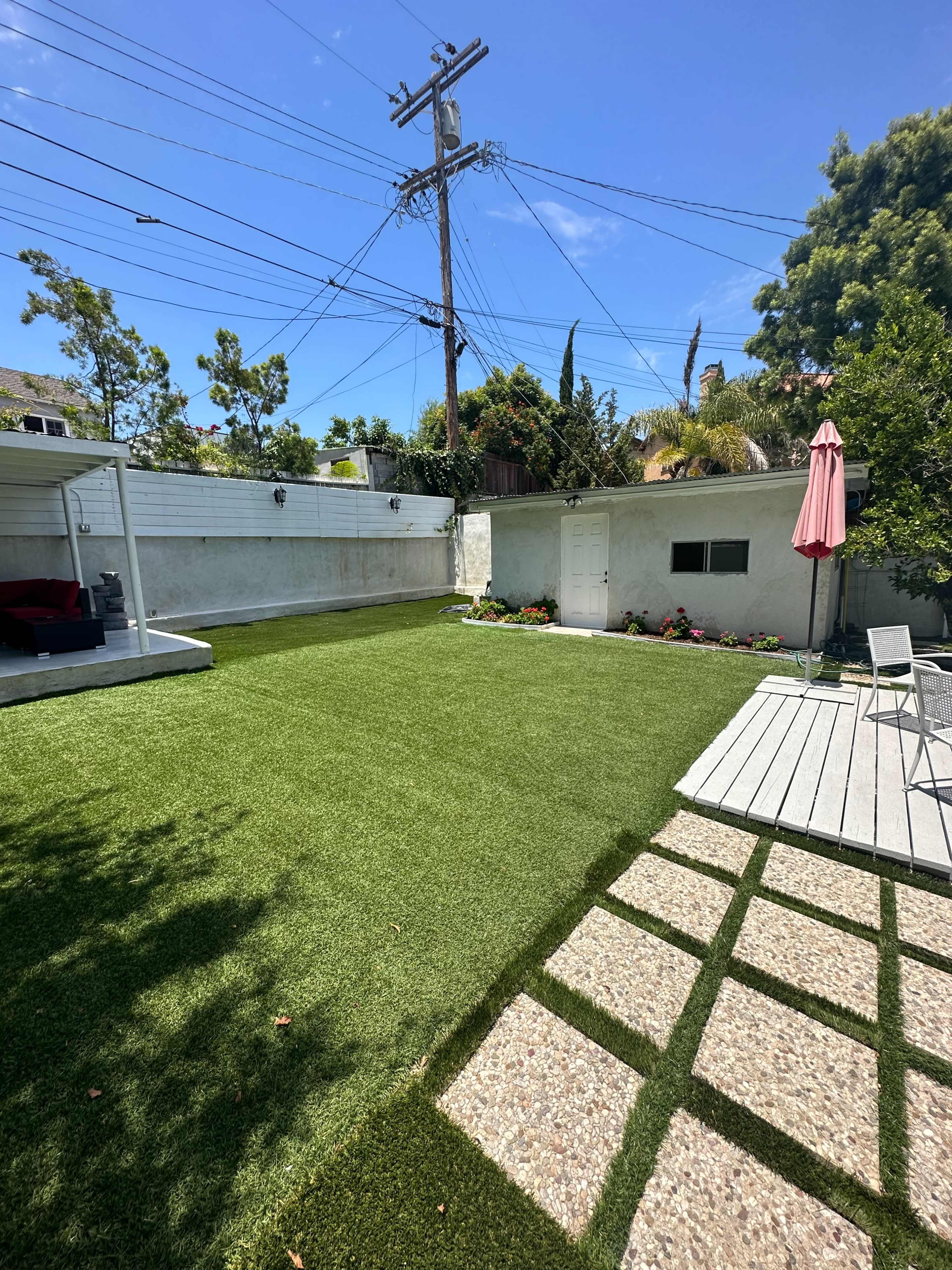 A landscaped backyard featuring artificial grass, a stone pathway, and a small patio with a pink umbrella.