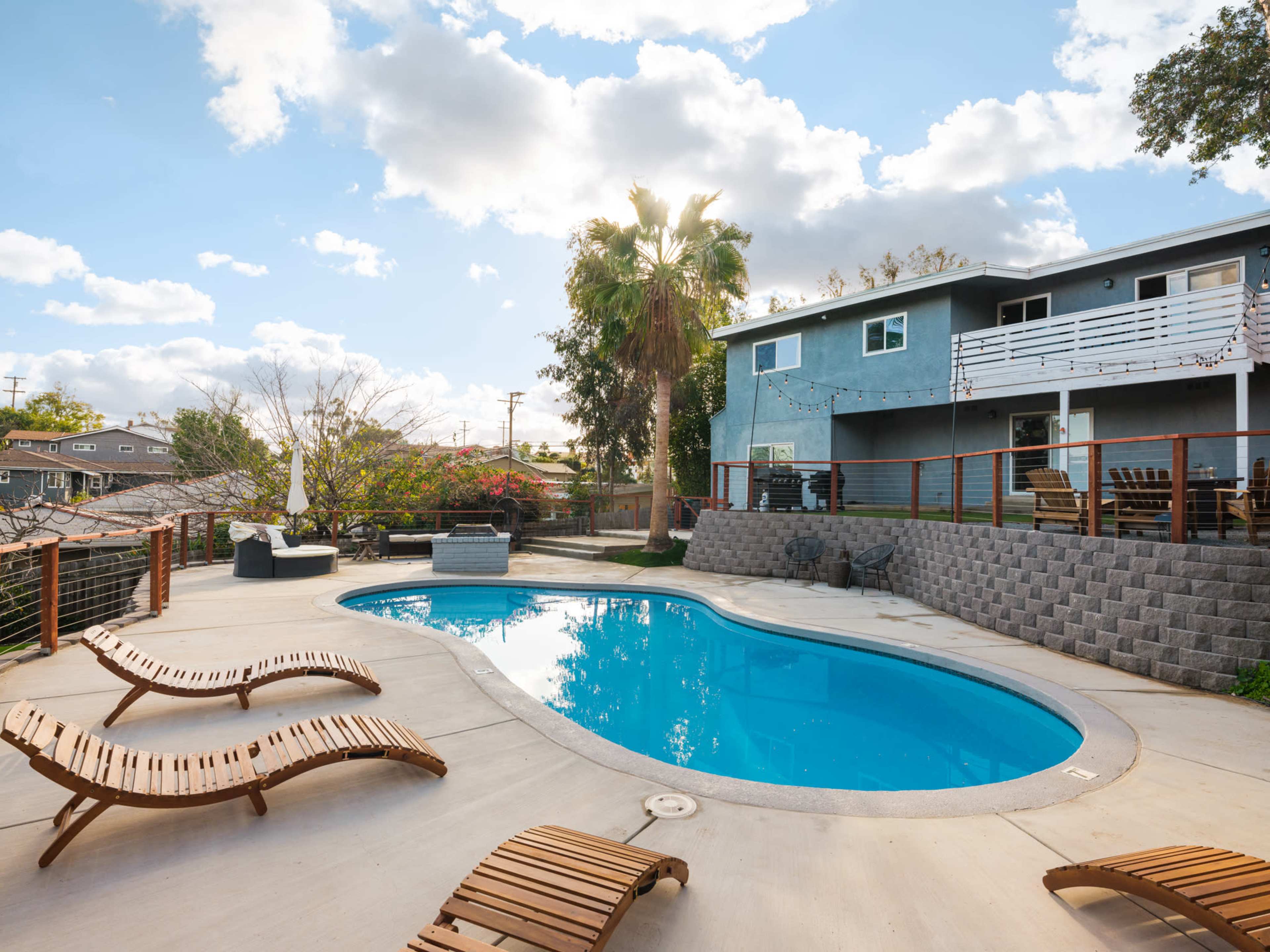 A rectangular swimming pool surrounded by wooden lounge chairs sits in a backyard with a two-story house and palm tree in the background.