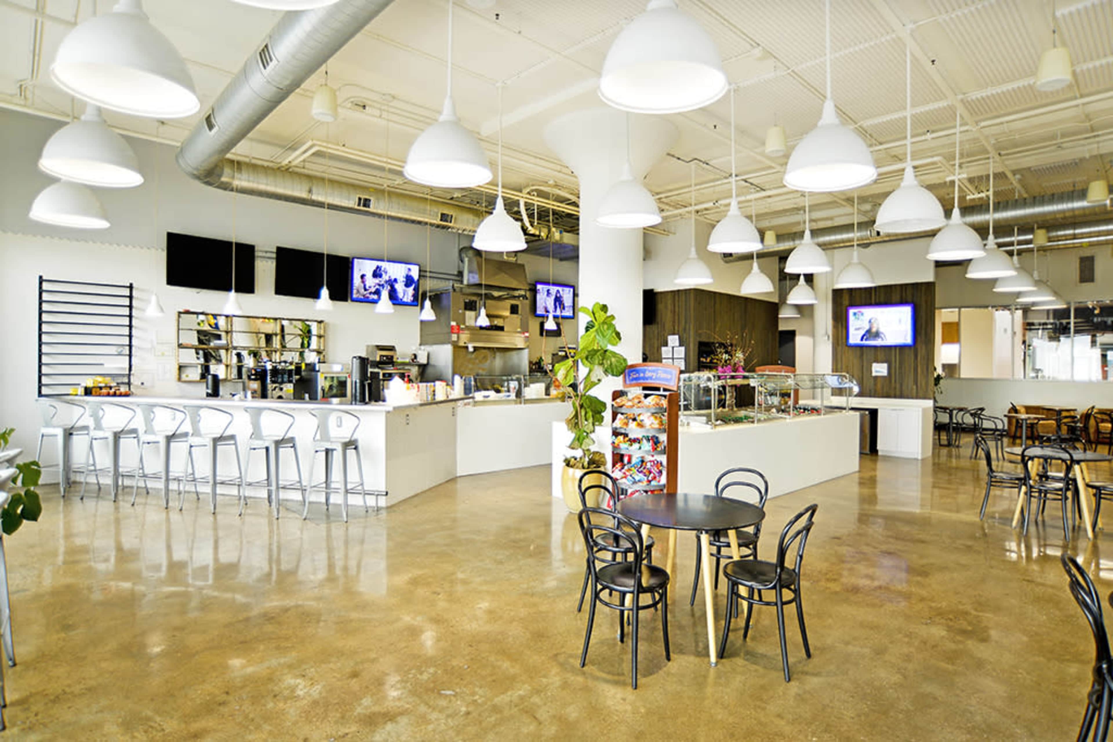 A spacious, modern café features a counter with a display case, bar seating, and tables arranged throughout the bright interior.