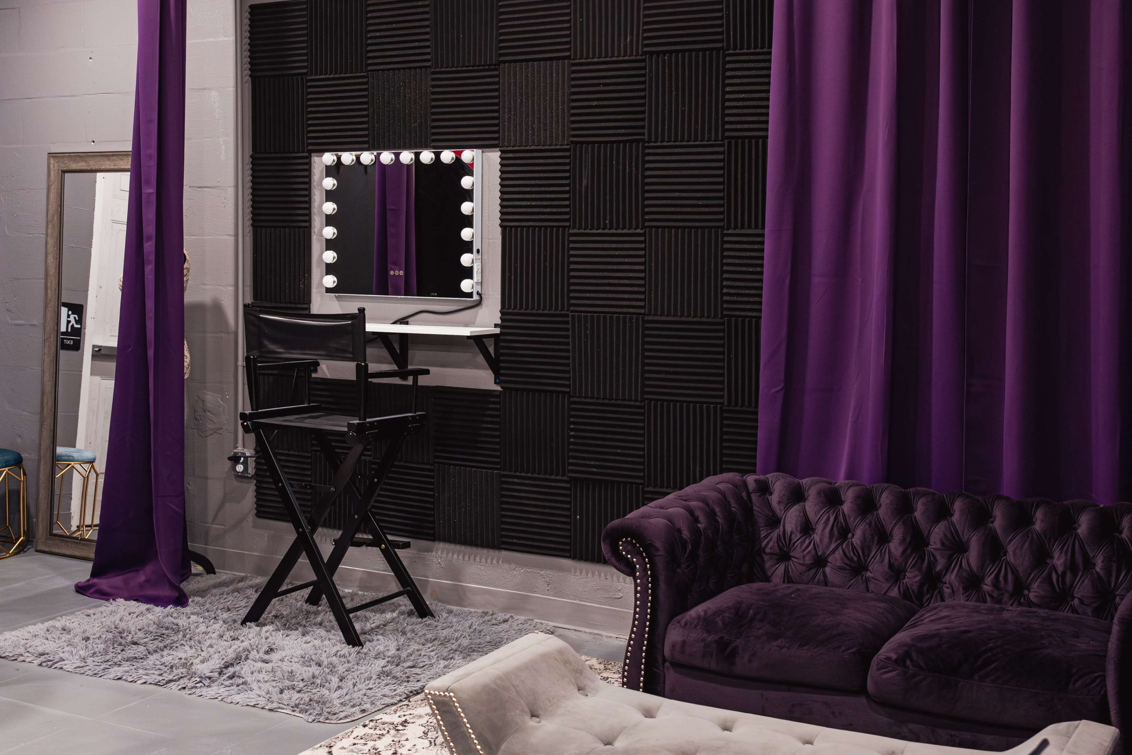 A cozy room with a purple sofa, a standing mirror, a makeup mirror with lights, and black acoustic panels on the wall.