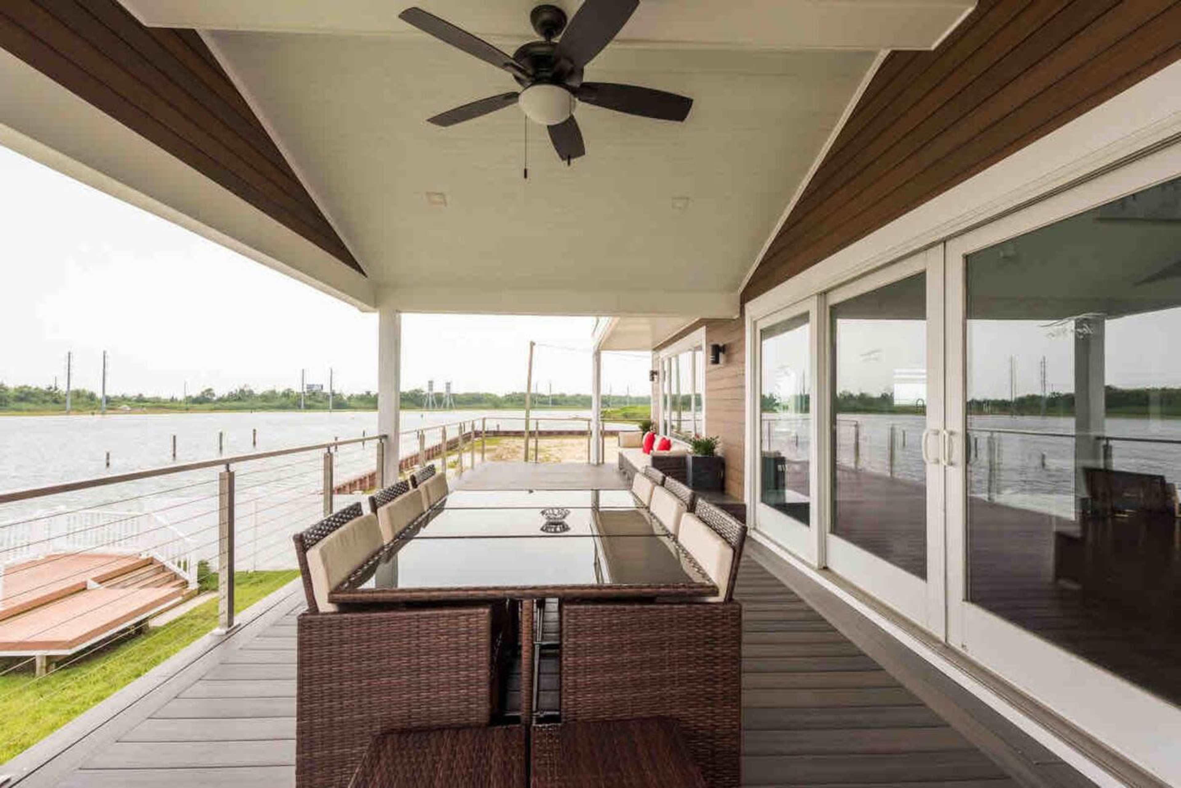 A covered outdoor dining area features a large table surrounded by wicker chairs, overlooking a body of water.