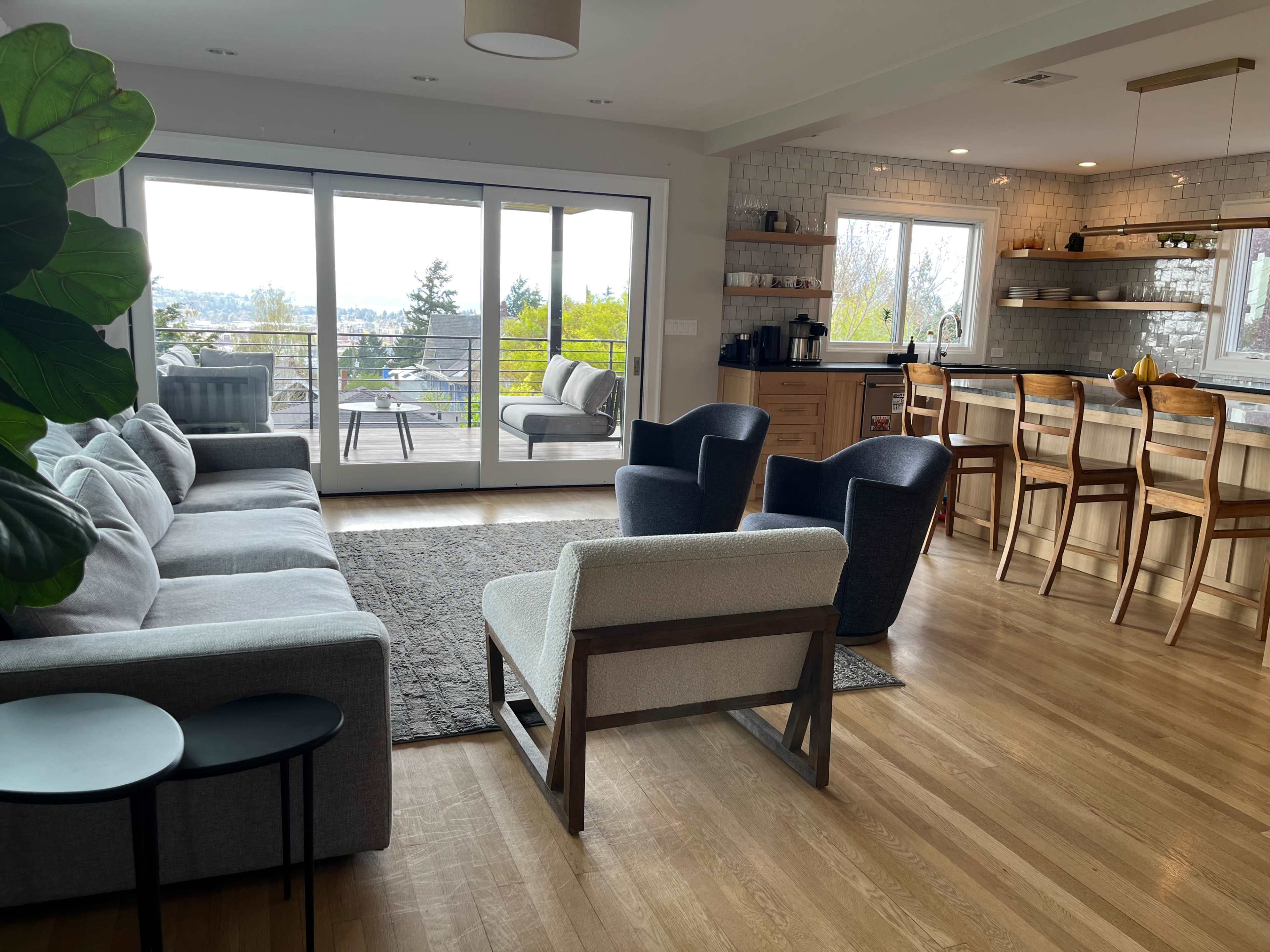 A modern living room features a light gray sectional sofa, two black chairs, and a small side table, with large windows opening to a deck and a view of trees and distant hills.